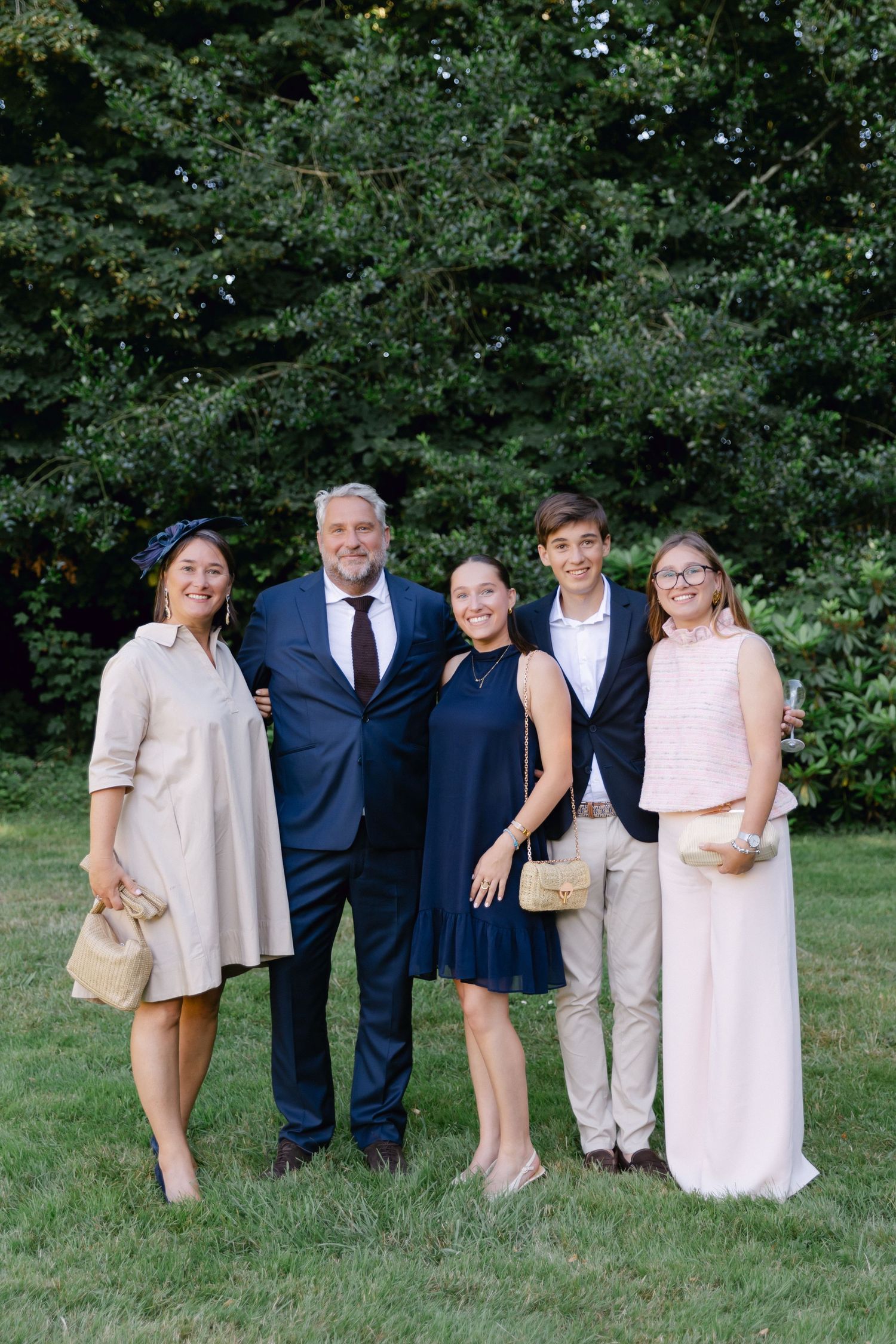 Une famille de cinq personnes pose à l'extérieur sur l'herbe verte, vêtue d'une tenue formelle, souriant ensemble avec des arbres luxuriants