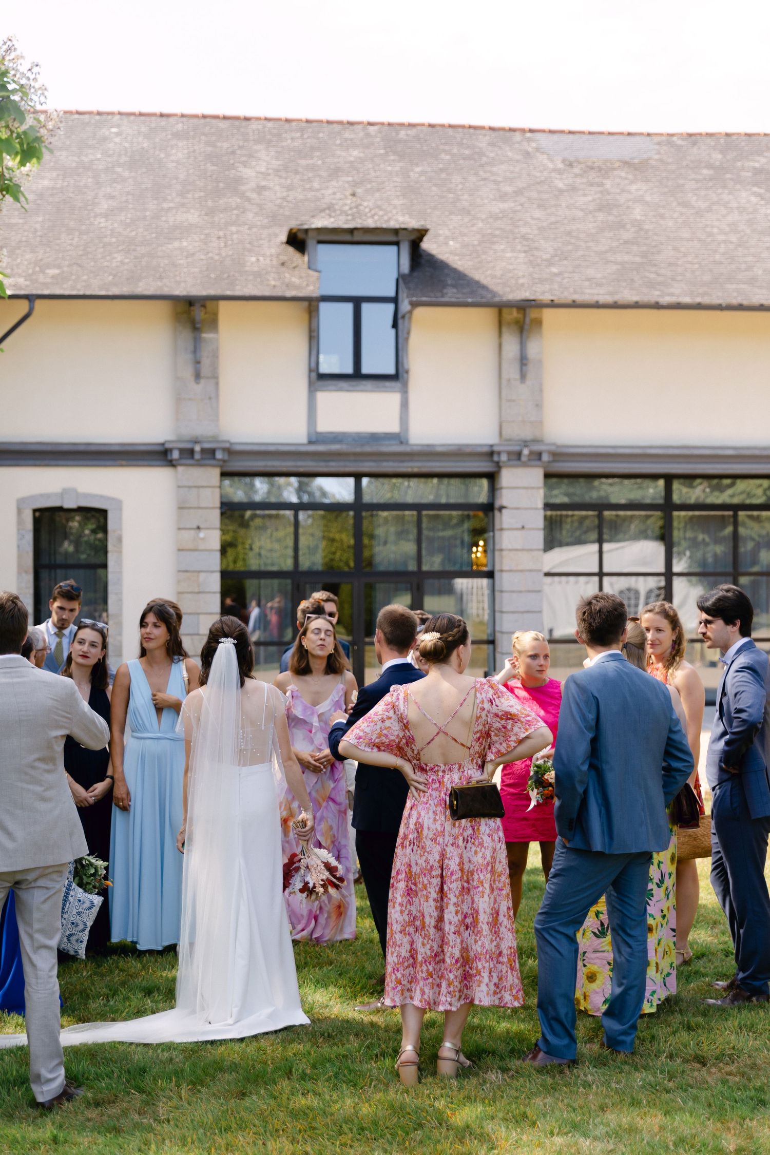 Les invités au mariage se rassemblent à l'extérieur sur une pelouse près d'un lieu élégant, avec une mariée en robe blanche et un voile visi