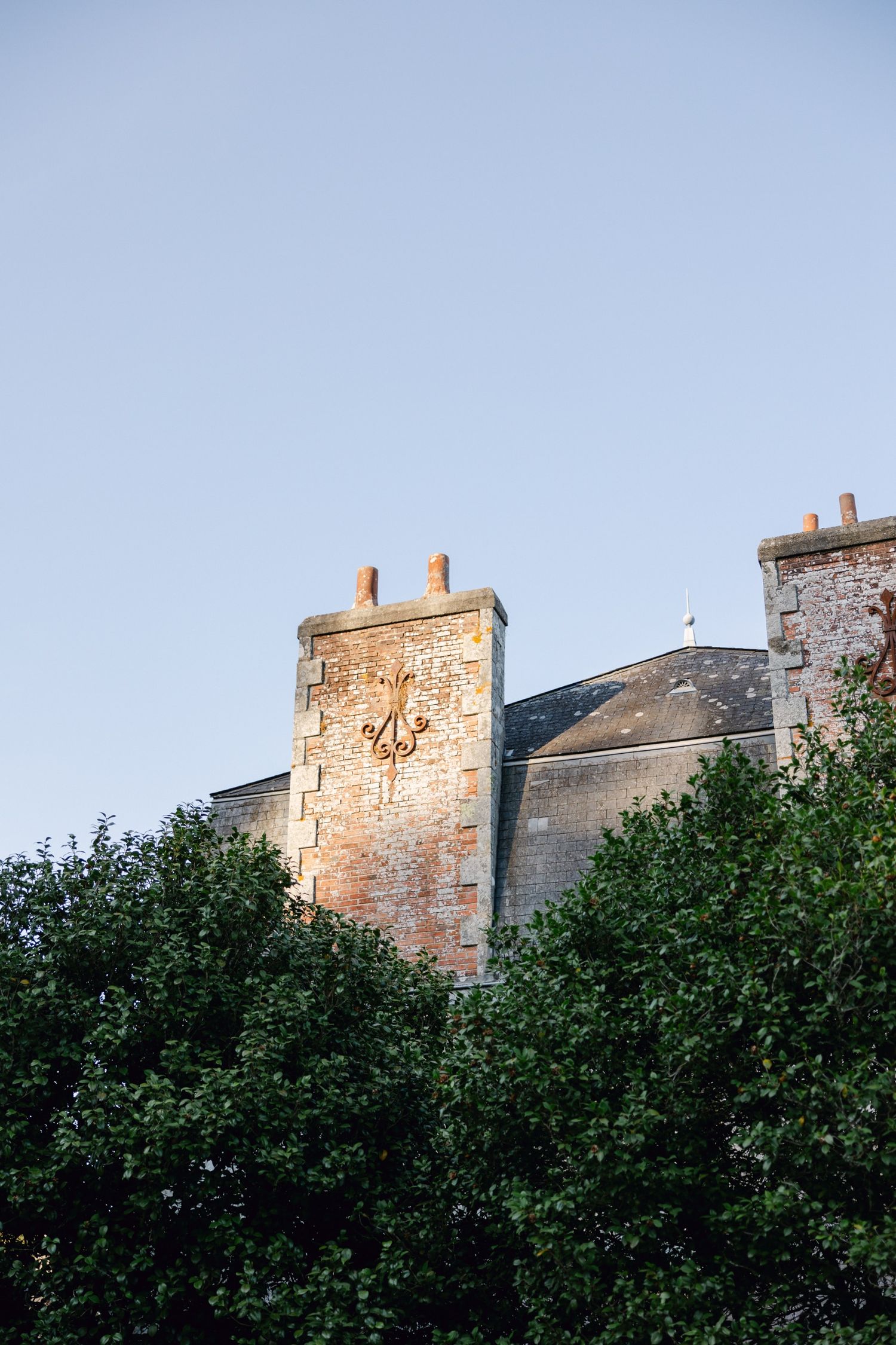 Historic brick castle tower with ornate golden emblem, framed by lush green trees against a clear blue sky.