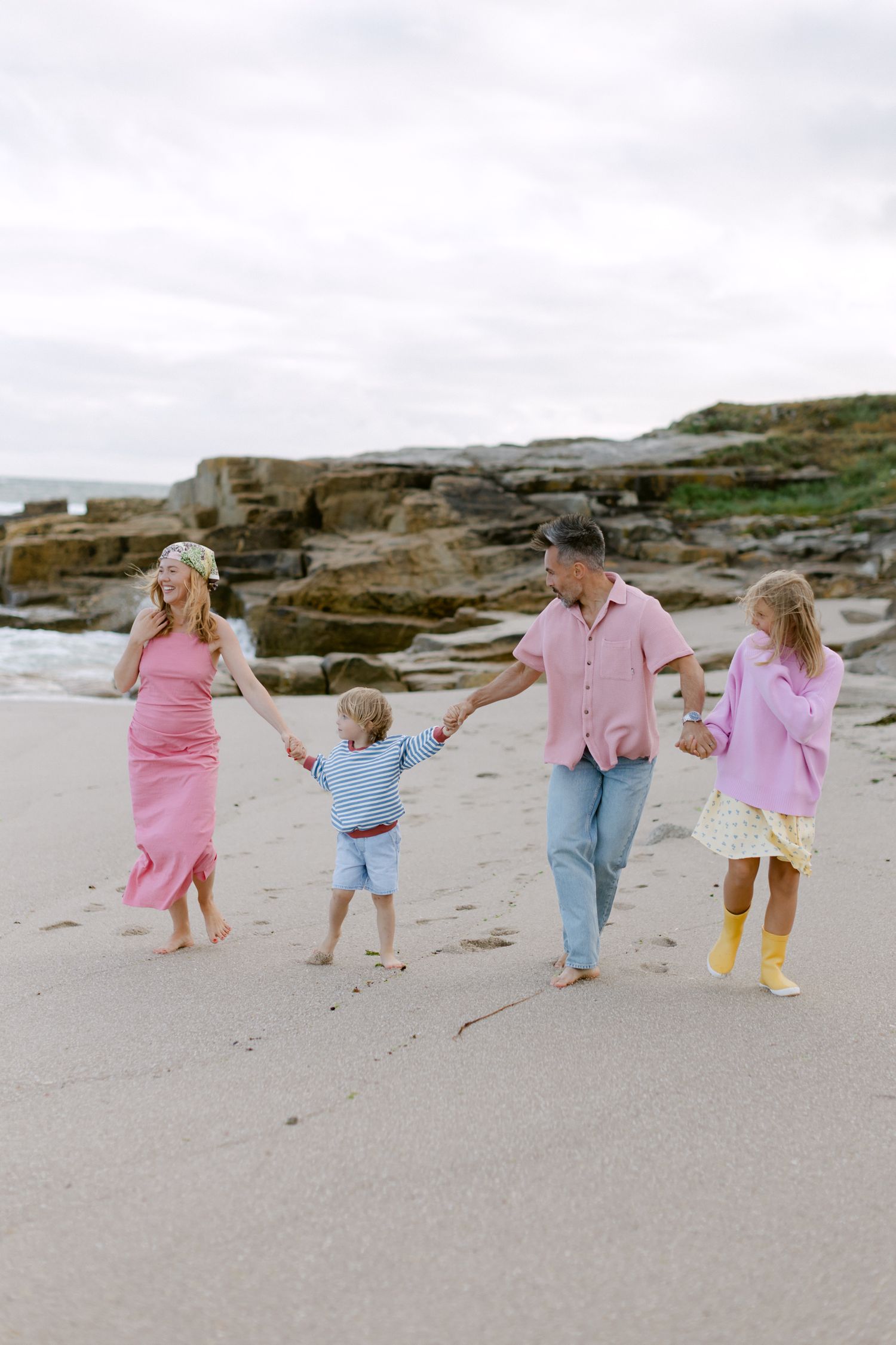 Family of four holding hands on a sandy beach, wearing pink and pastel outfits, with rocky coastline in the background.