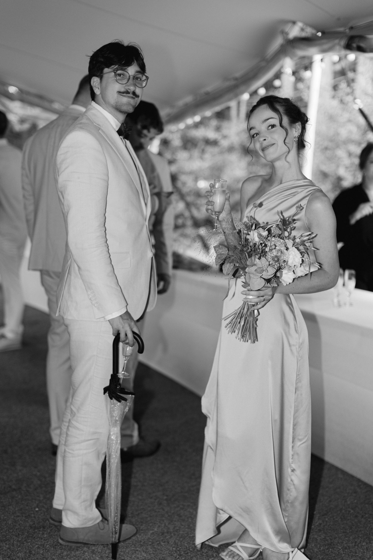 Photo en noir et blanc d'invités de mariage élégamment habillés posant avec un bouquet sous la tente de réception.