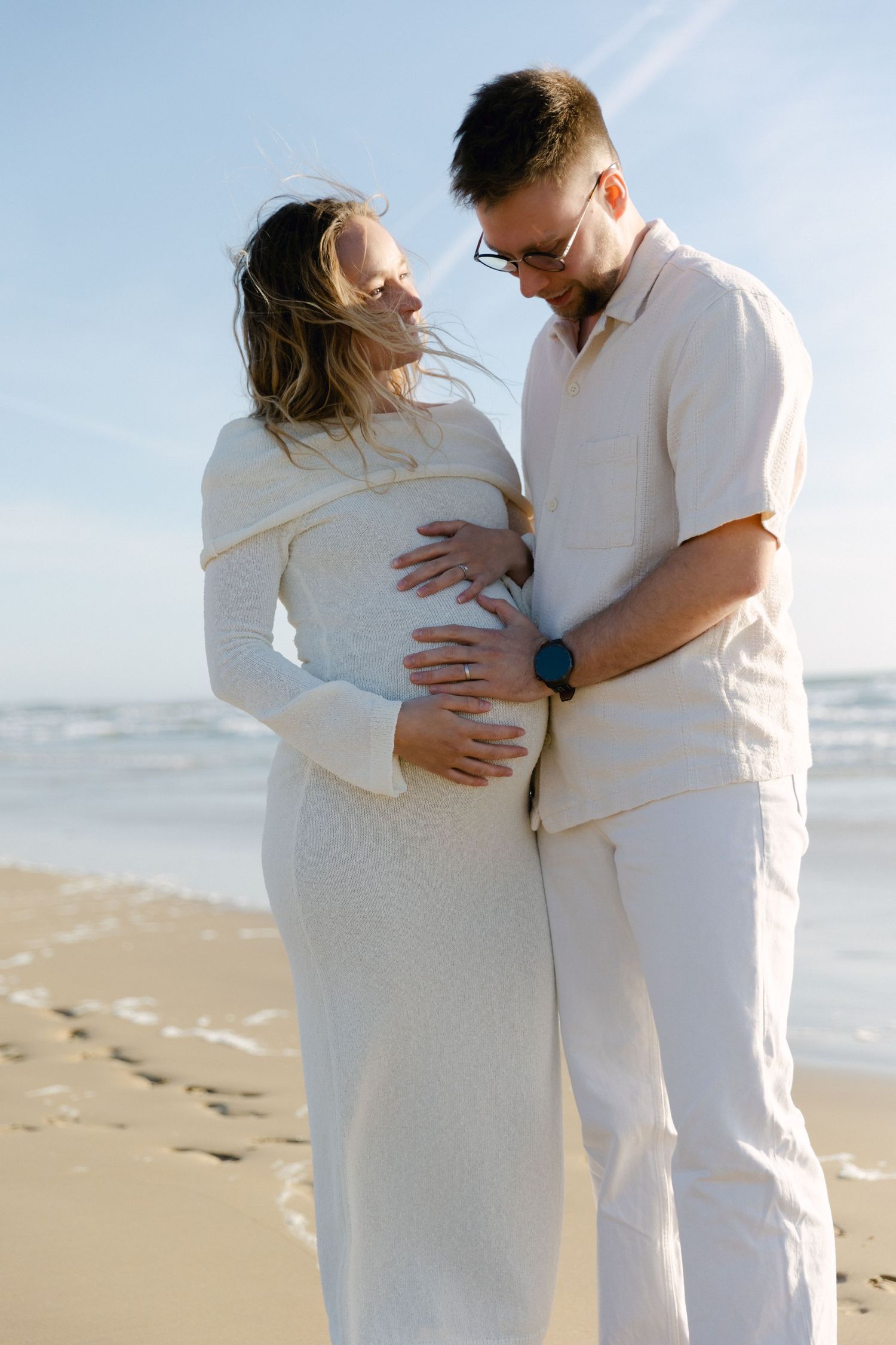Couple s'embrassant sur la plage, les mains sur le ventre de femme enceinte, portant des tenues crème assorties avec une toile de fond océan