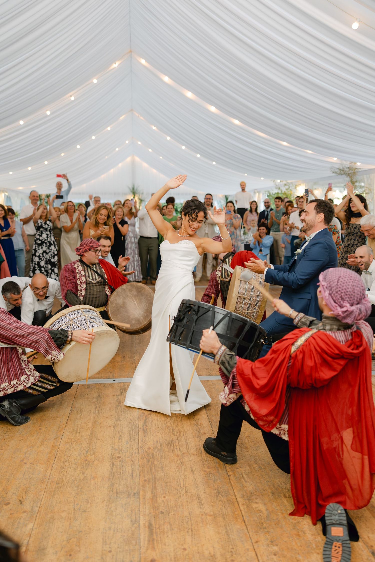 Bride dances joyfully with live drummers in traditional attire at a marquee wedding reception with guests cheering.