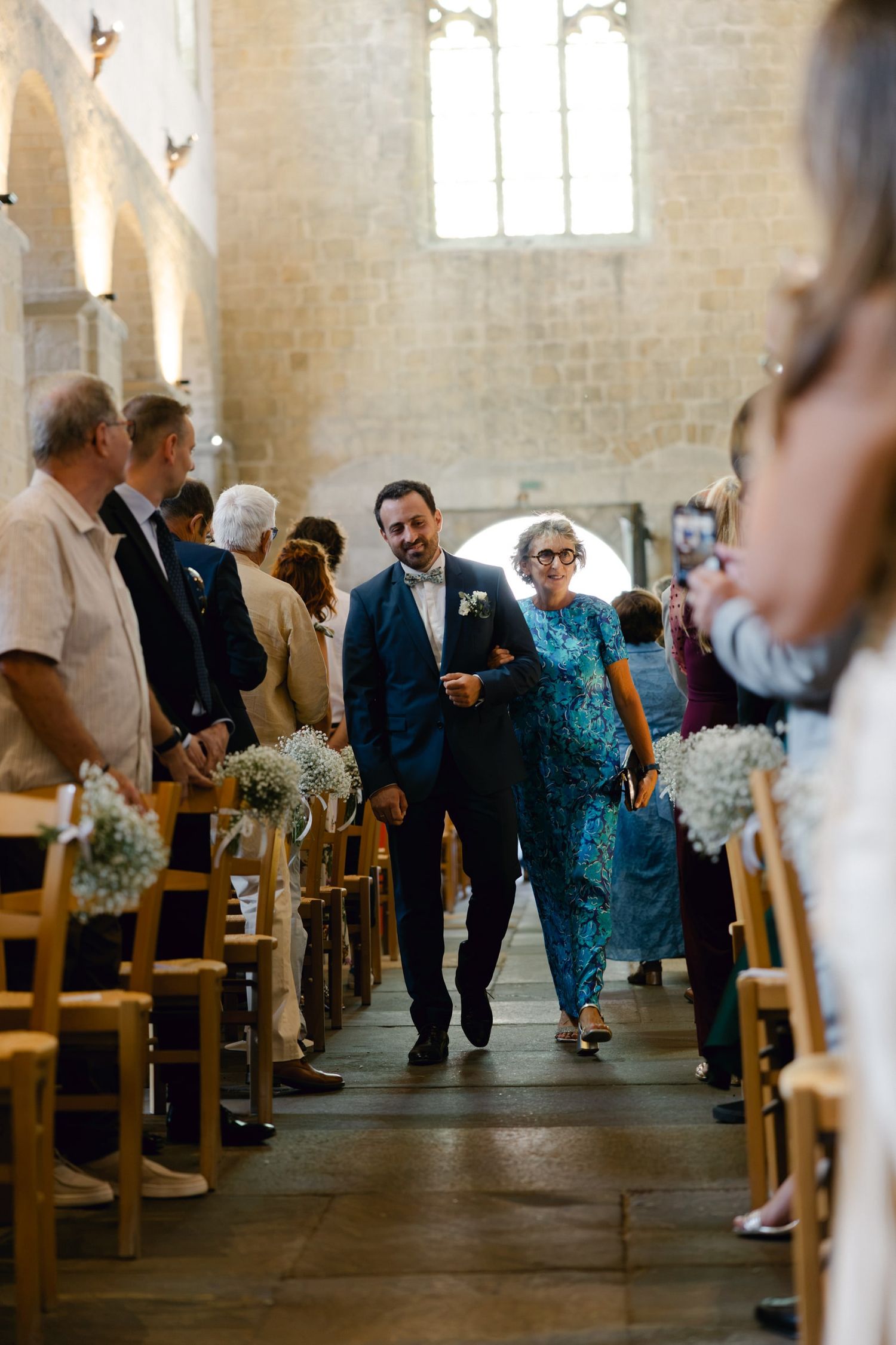 Groom walks down church aisle with mother during wedding ceremony, guests standing in pews decorated with floral arrangements.