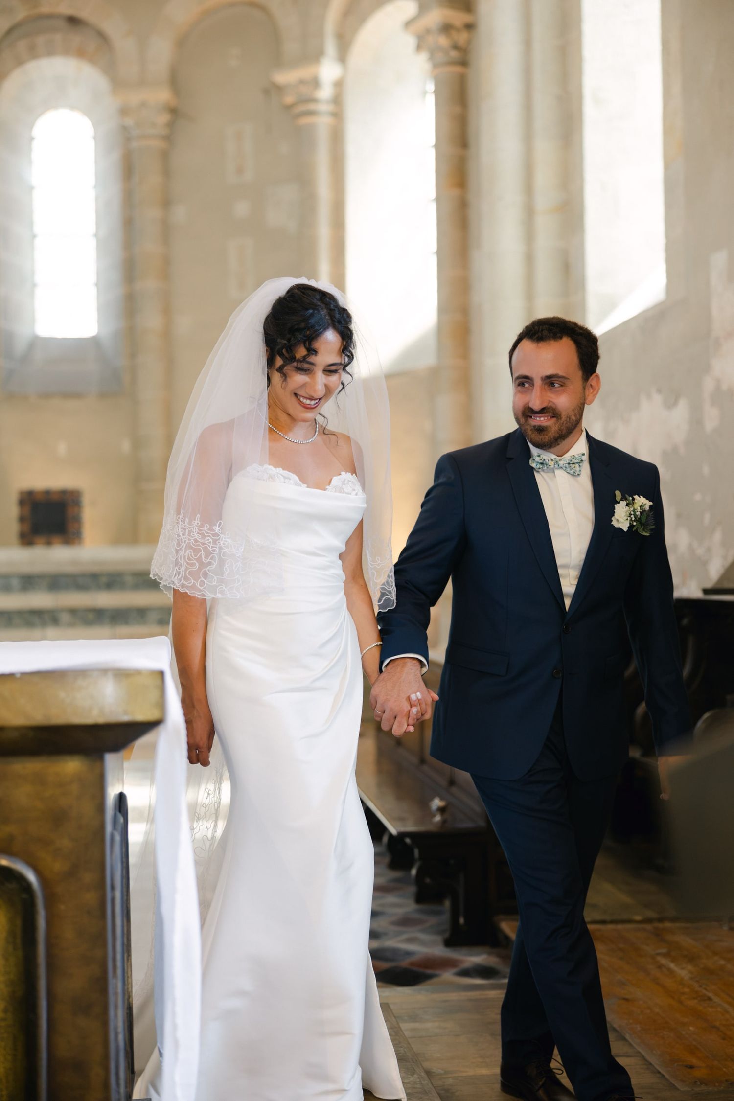 Happy newlyweds walking hand in hand down the aisle of a sunlit church, bride in white gown and veil.