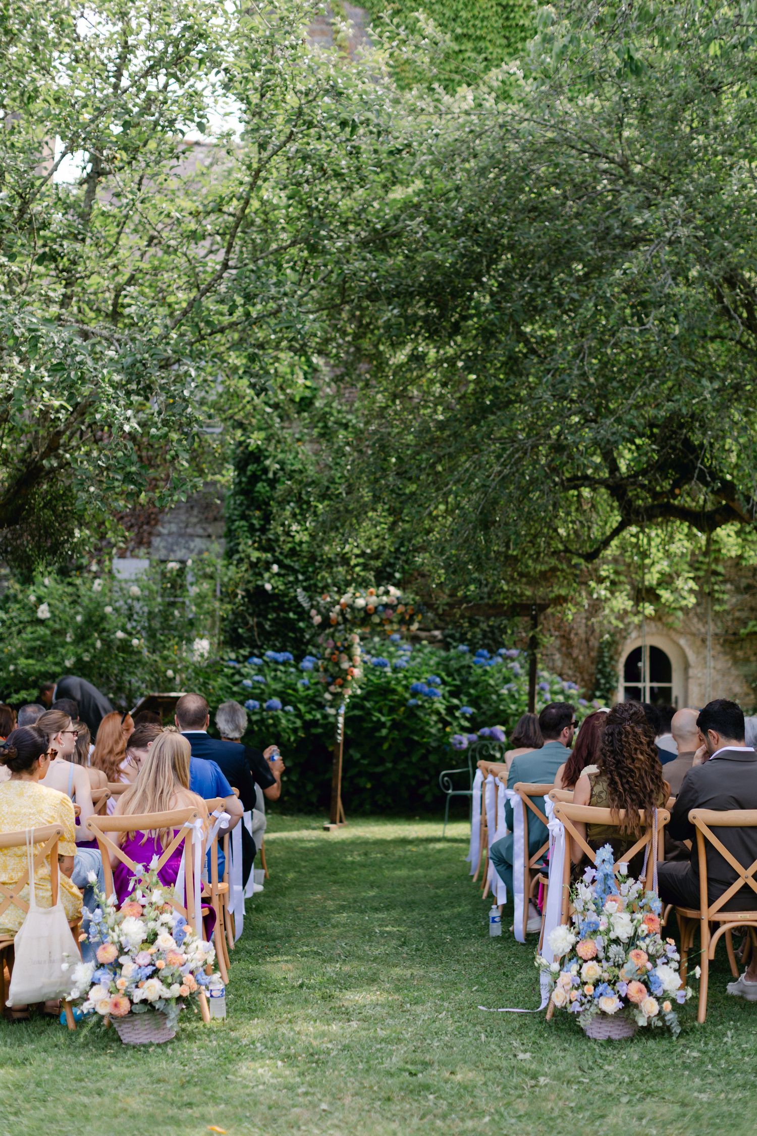 Élégante allée de cérémonie de mariage en plein air bordée de chaises en bois et d'arrangements floraux sous des arbres verts.