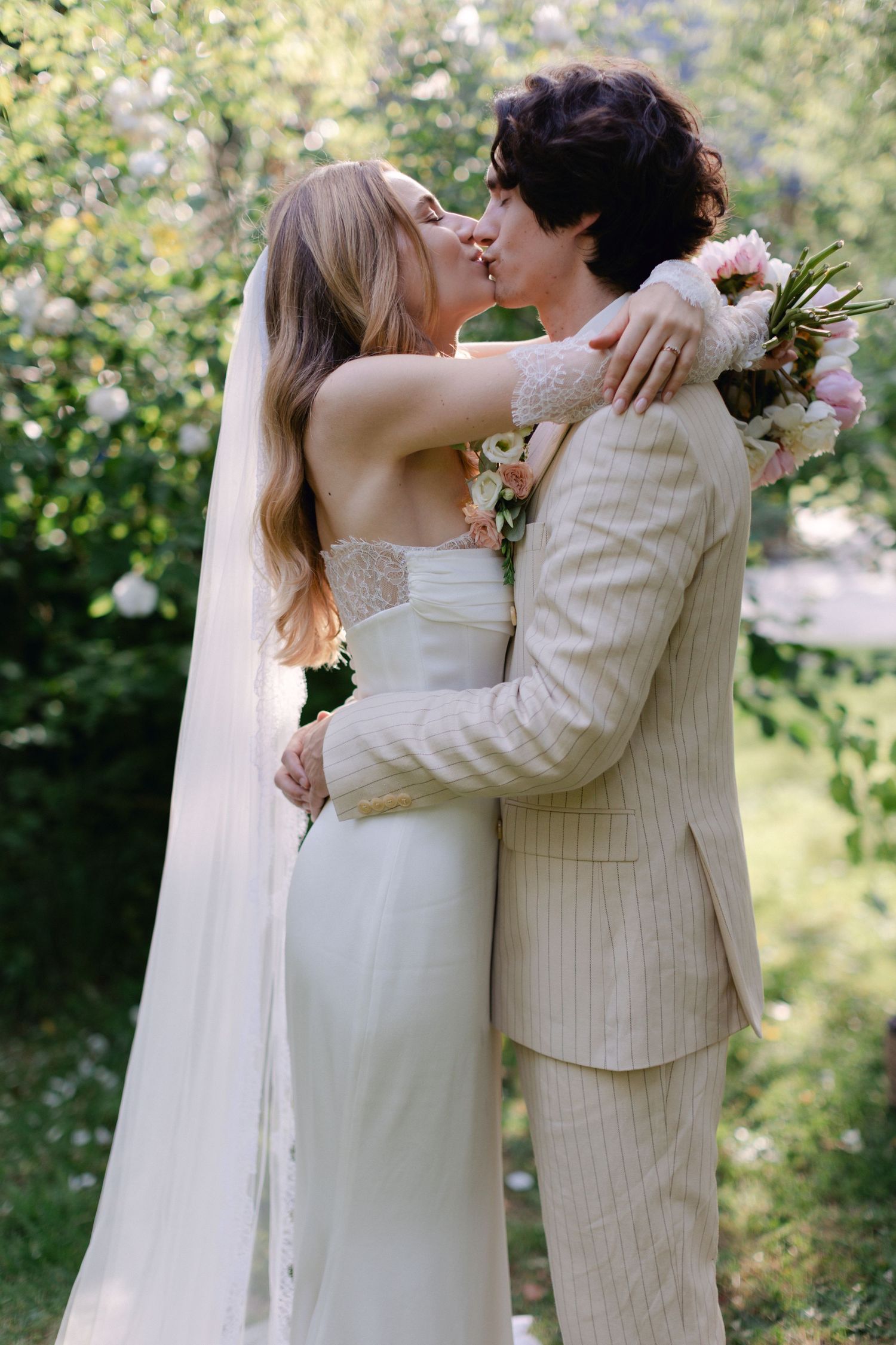 La mariée et le marié partagent un baiser à l'extérieur, elle dans une robe blanche sans bretelles avec voile, lui dans un costume à rayures