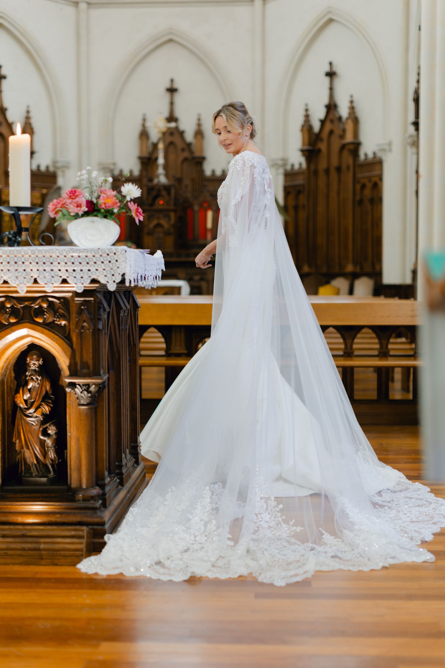 Une mariée élégante dans une robe de mariée en dentelle blanche ajustée avec un voile de cape transparent pose à l'intérieur d'une église or