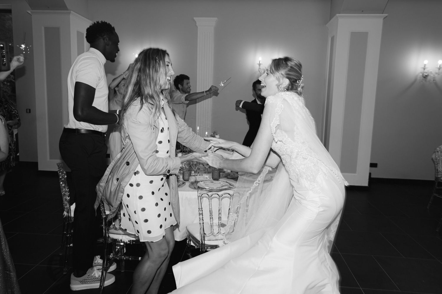 Photo en noir et blanc d'une mariée en robe de mariée dansant avec des invités lors d'une réception de mariage animée.