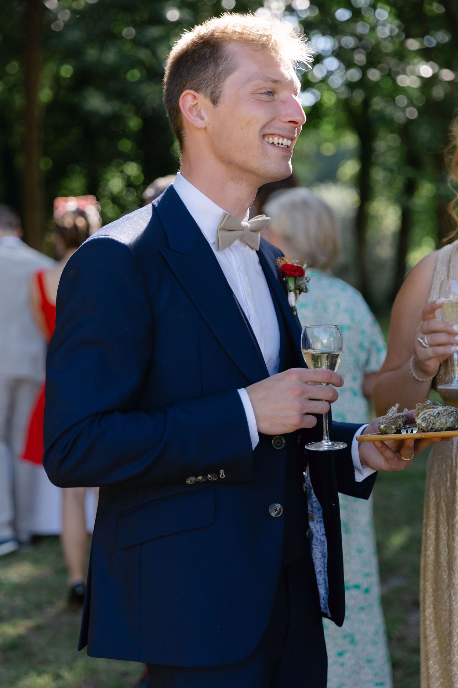 Marié souriant en costume bleu marine tenant une coupe de champagne lors d'une réception de mariage en plein air.