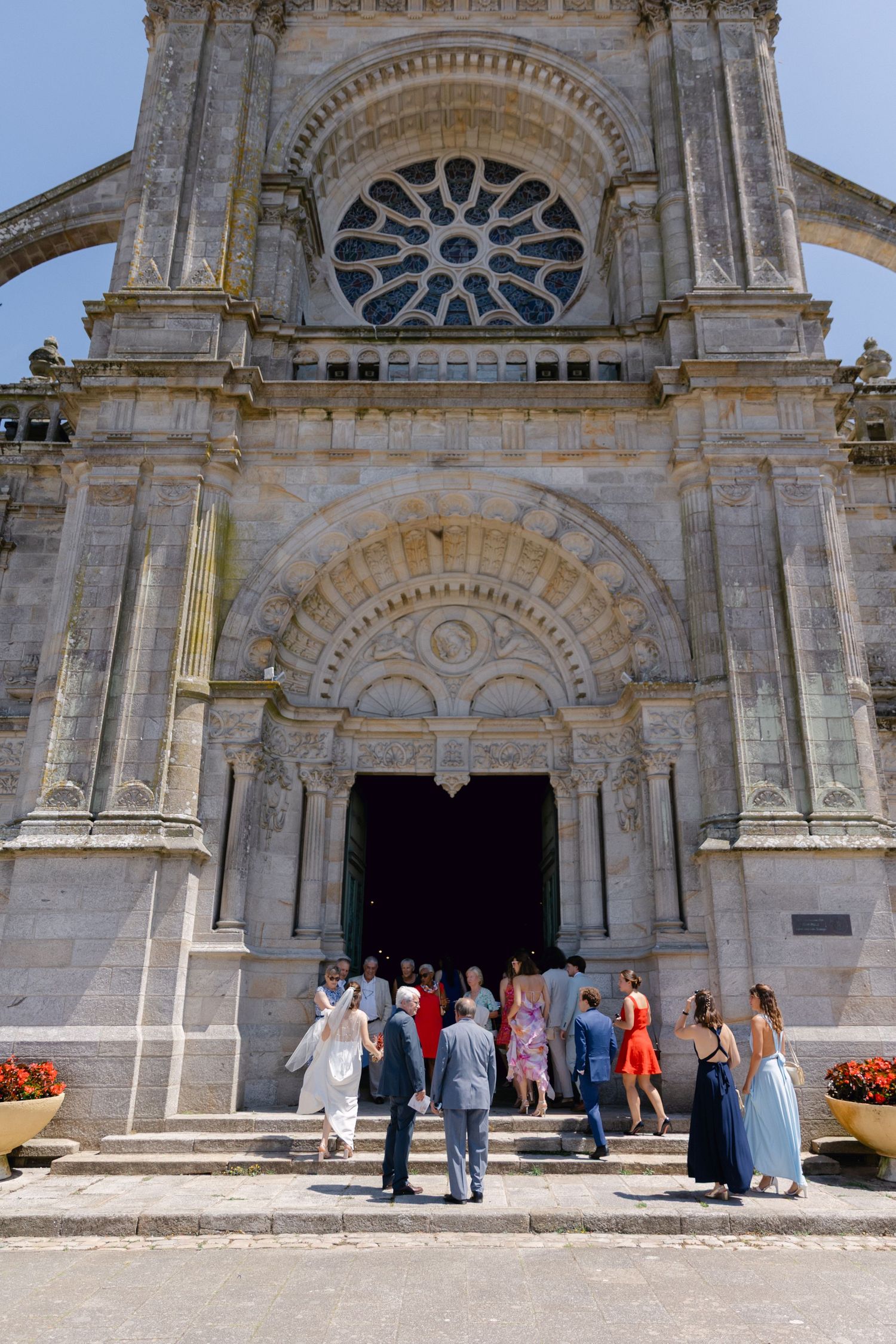 Élégants invités de mariage entrant dans une cathédrale en pierre ornée avec une rosette et une porte arquée par une journée ensoleillée.