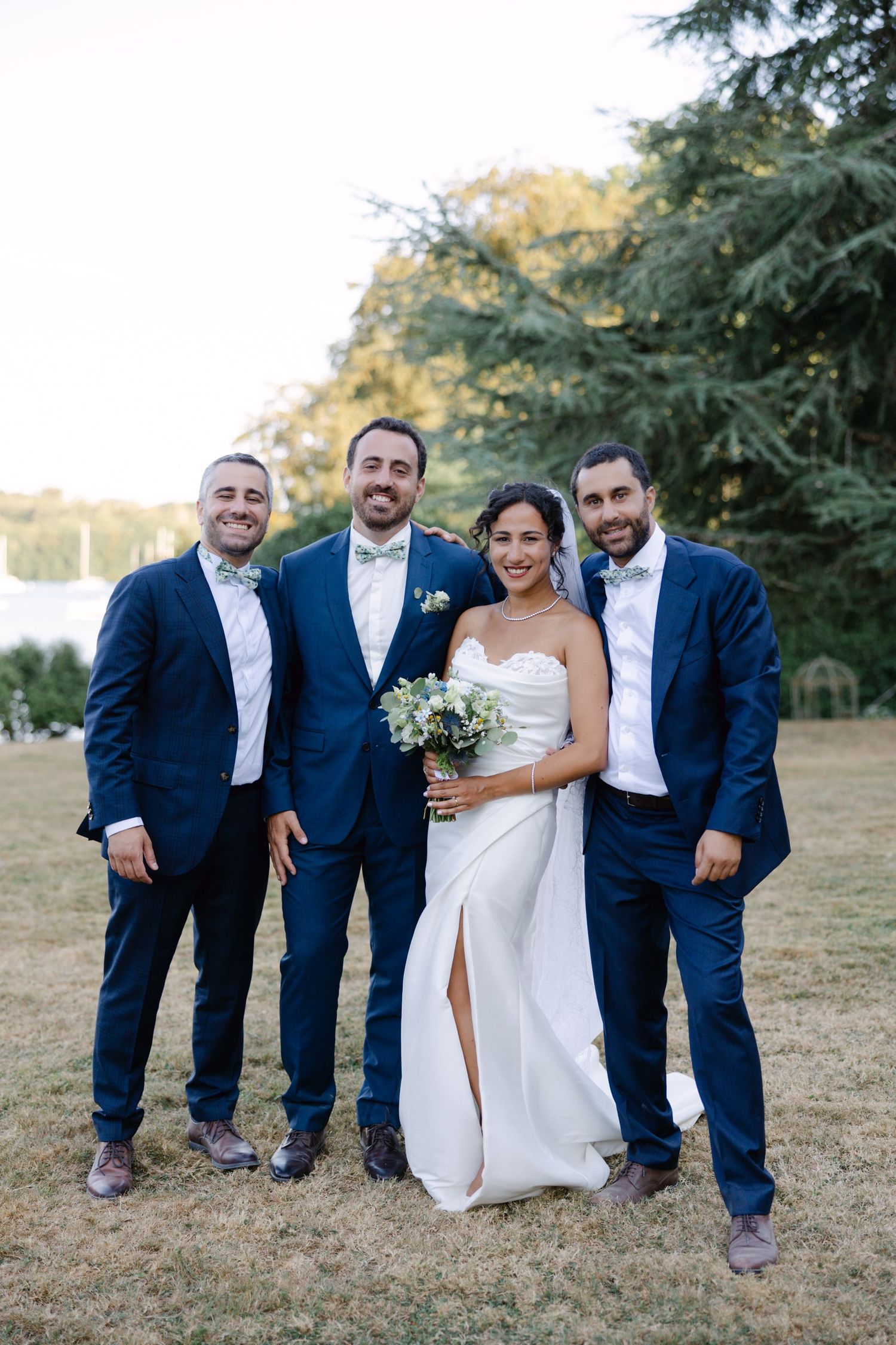 Bride in white gown with bouquet poses with three groomsmen in navy suits outdoors.