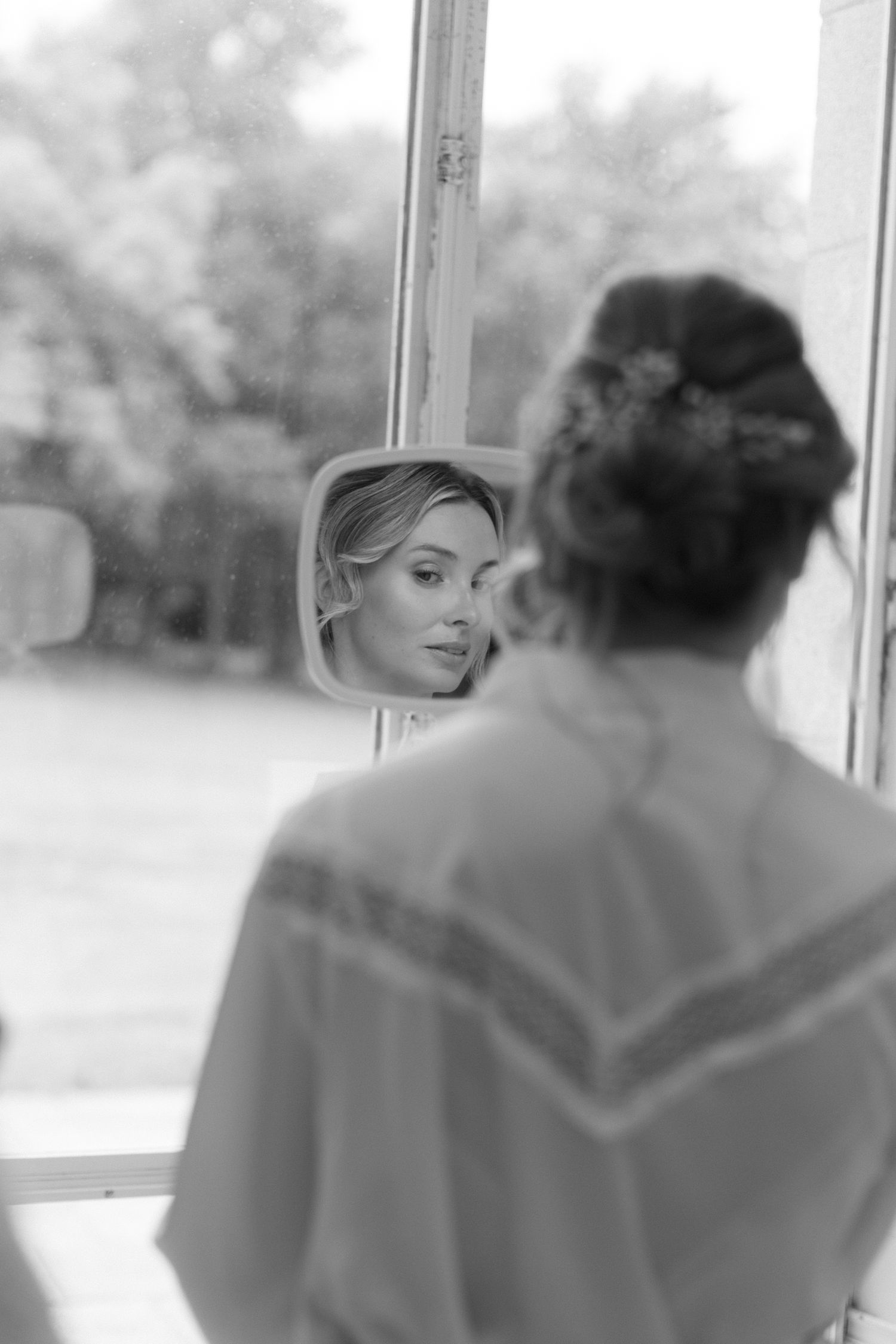 Portrait en noir et blanc d'une mariée vérifiant son reflet dans un miroir de fenêtre avec une élégante coiffure.
