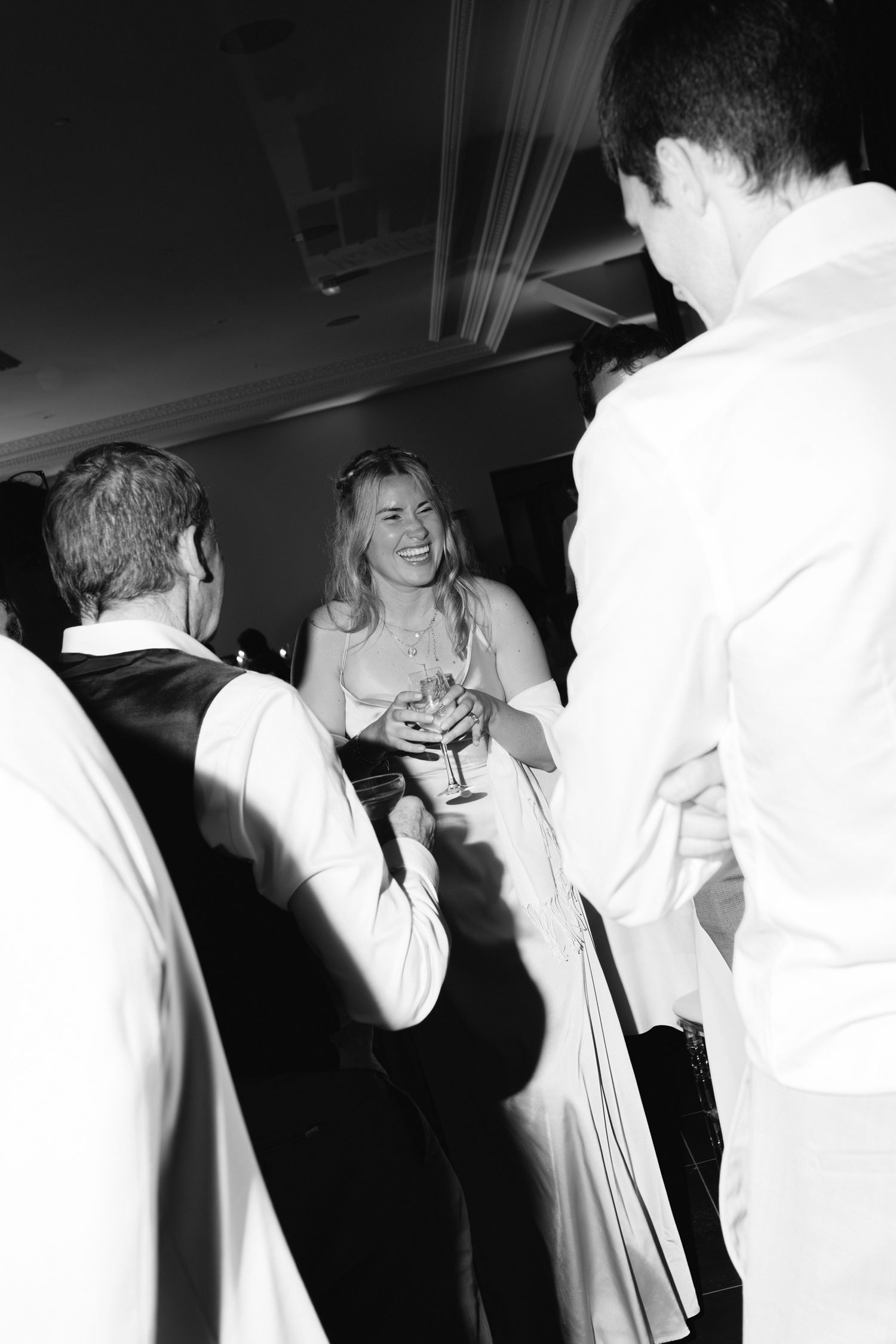 Demoiselle d'honneur riante en robe blanche tenant une boisson entourée d'invités lors d'une réception de mariage animée en noir et blanc.
