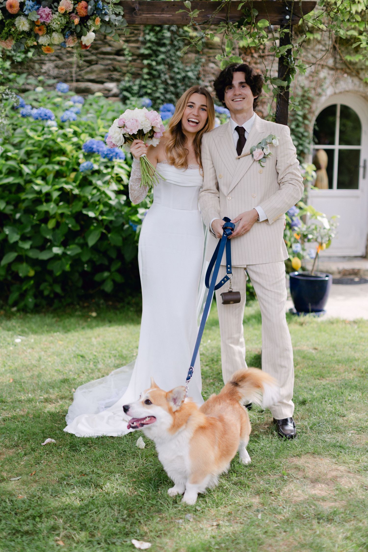 Mariée et marié posant avec un corgi en laisse dans un jardin luxuriant, la mariée tenant un bouquet rose dans une robe blanche.