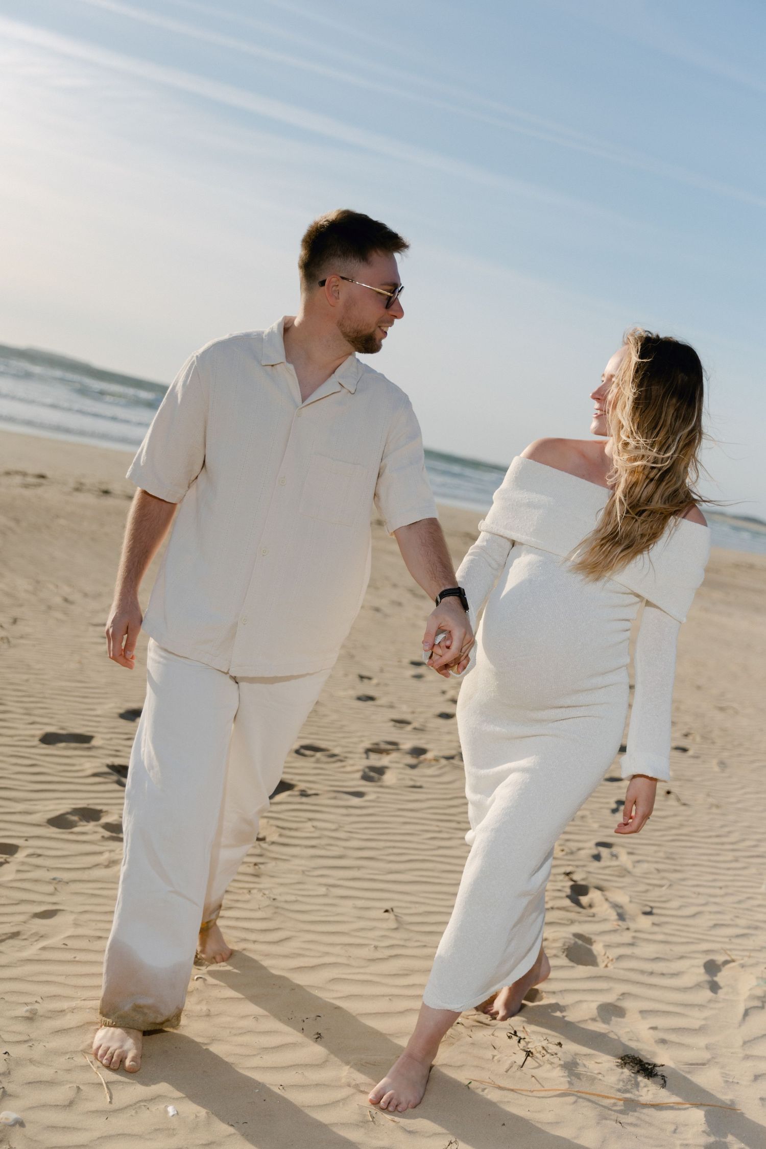Couple en tenue blanche se tenant la main et marchant ensemble sur une plage de sable près de l'océan.