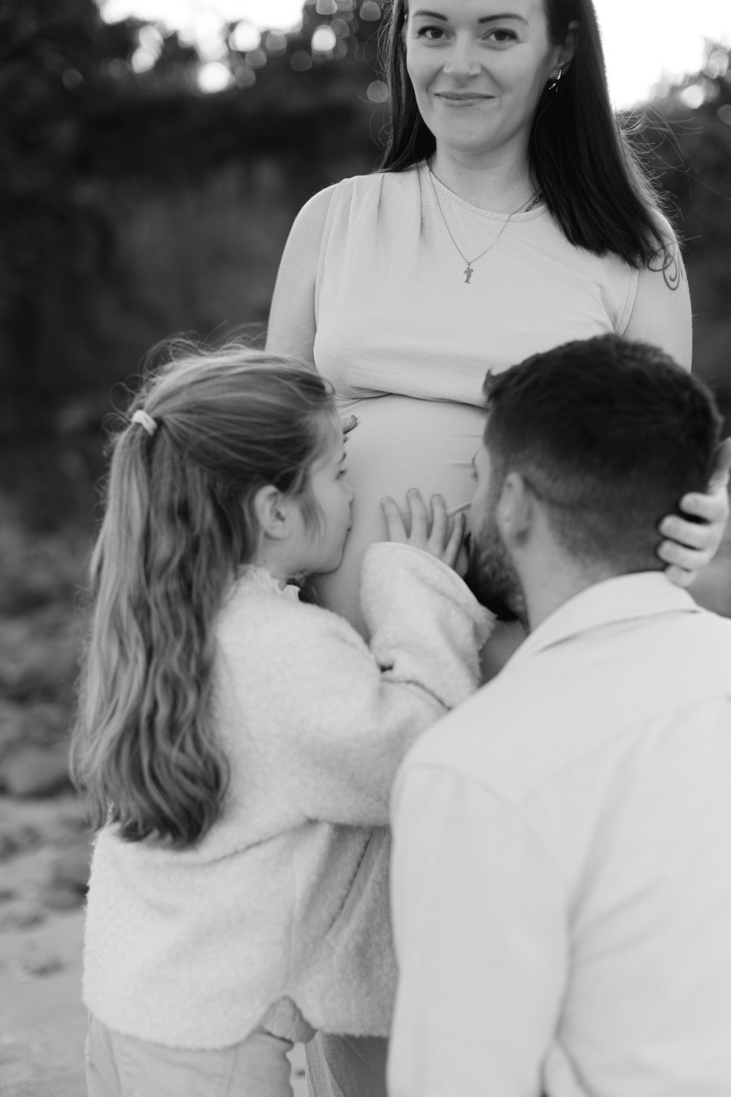 Photo en noir et blanc d'un enfant et d'un père embrassant la bosse d'une mère enceinte.