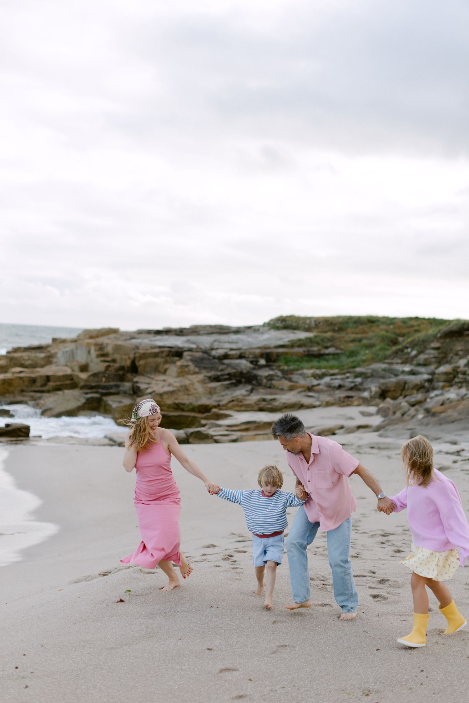 Famille de quatre personnes marchant main dans la main sur une plage rocheuse, portant des tenues roses et pastel par une journée nuageuse.
