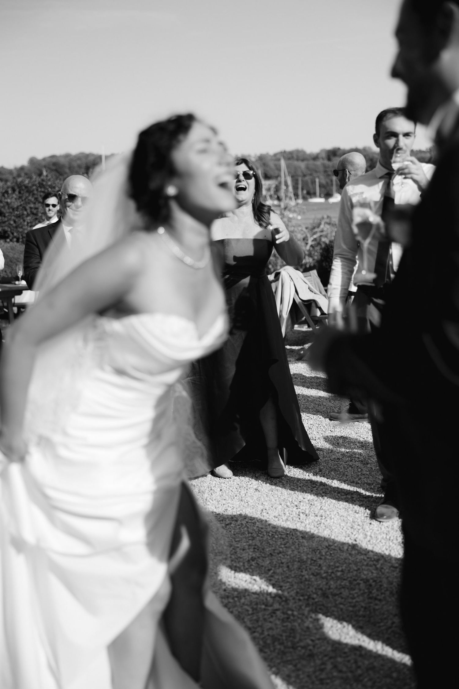 Black and white photo of a joyful bride in a strapless gown laughing outdoors at a wedding reception.