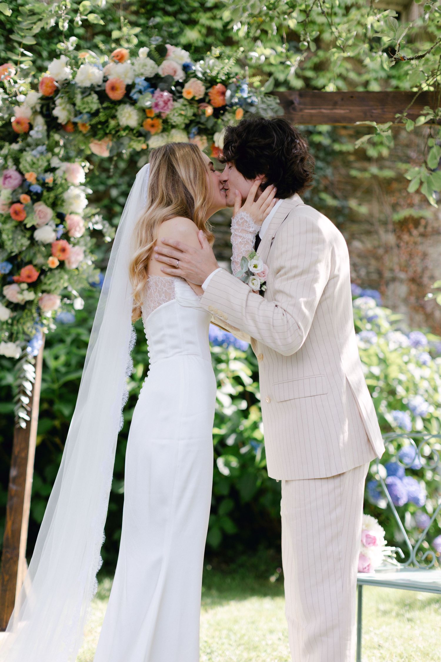 Les jeunes mariés partagent un premier baiser romantique sous une arche florale colorée lors d'un mariage dans un jardin en plein air.