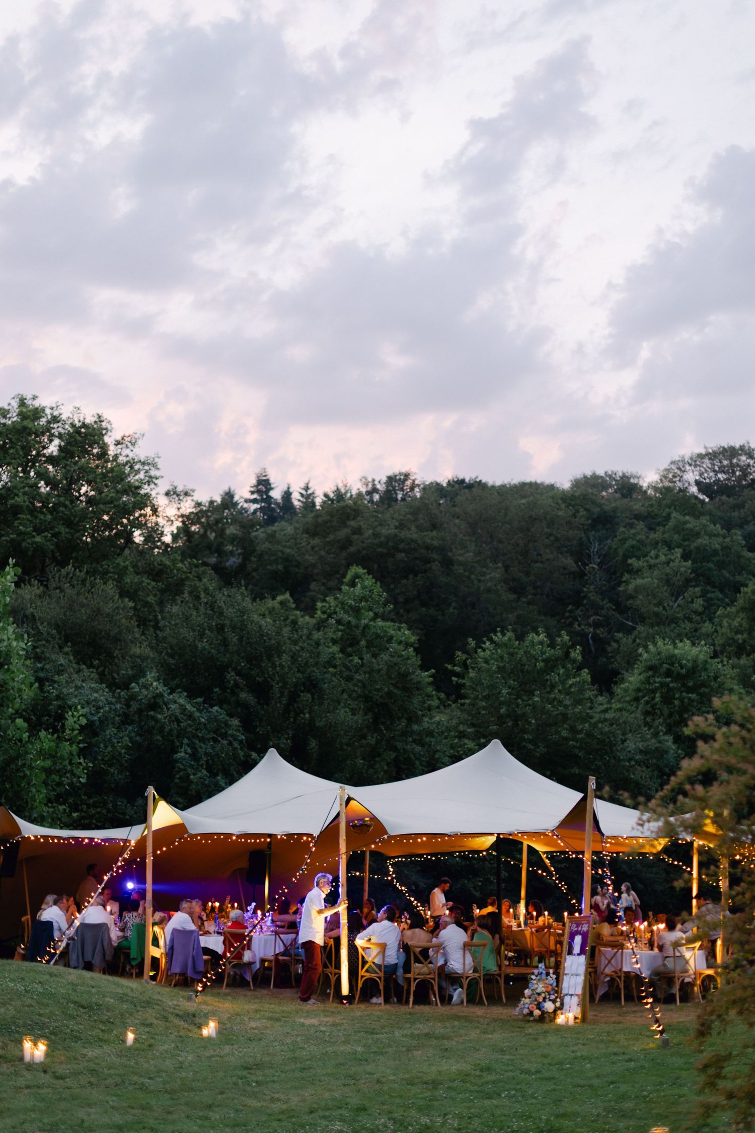 Réception de mariage magique en plein air sous des tentes extensibles éclairées au crépuscule entourées d'arbres verts luxuriants.