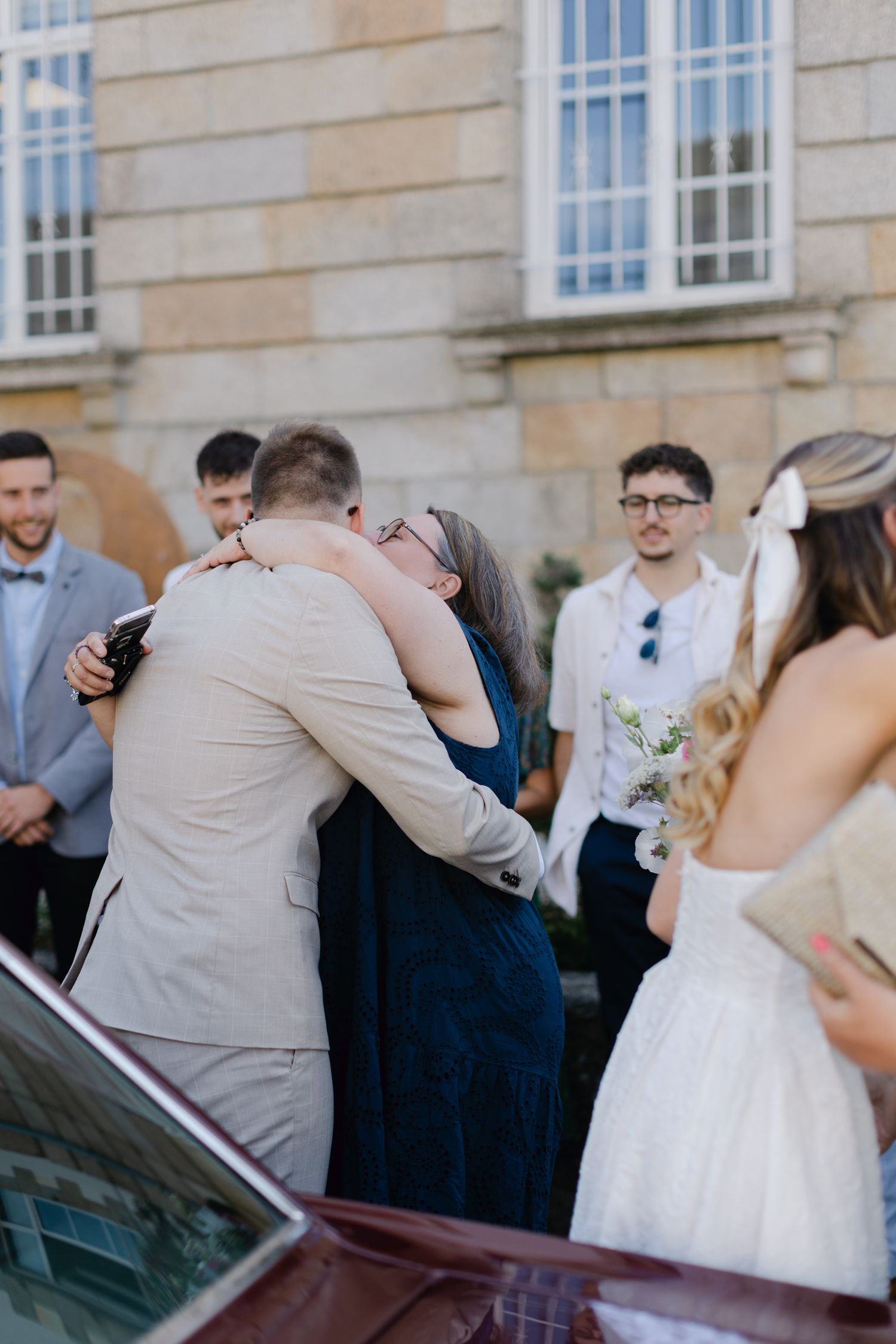 Le marié en costume beige partage une étreinte chaleureuse avec l'invité en robe marine à l'extérieur du lieu du mariage.