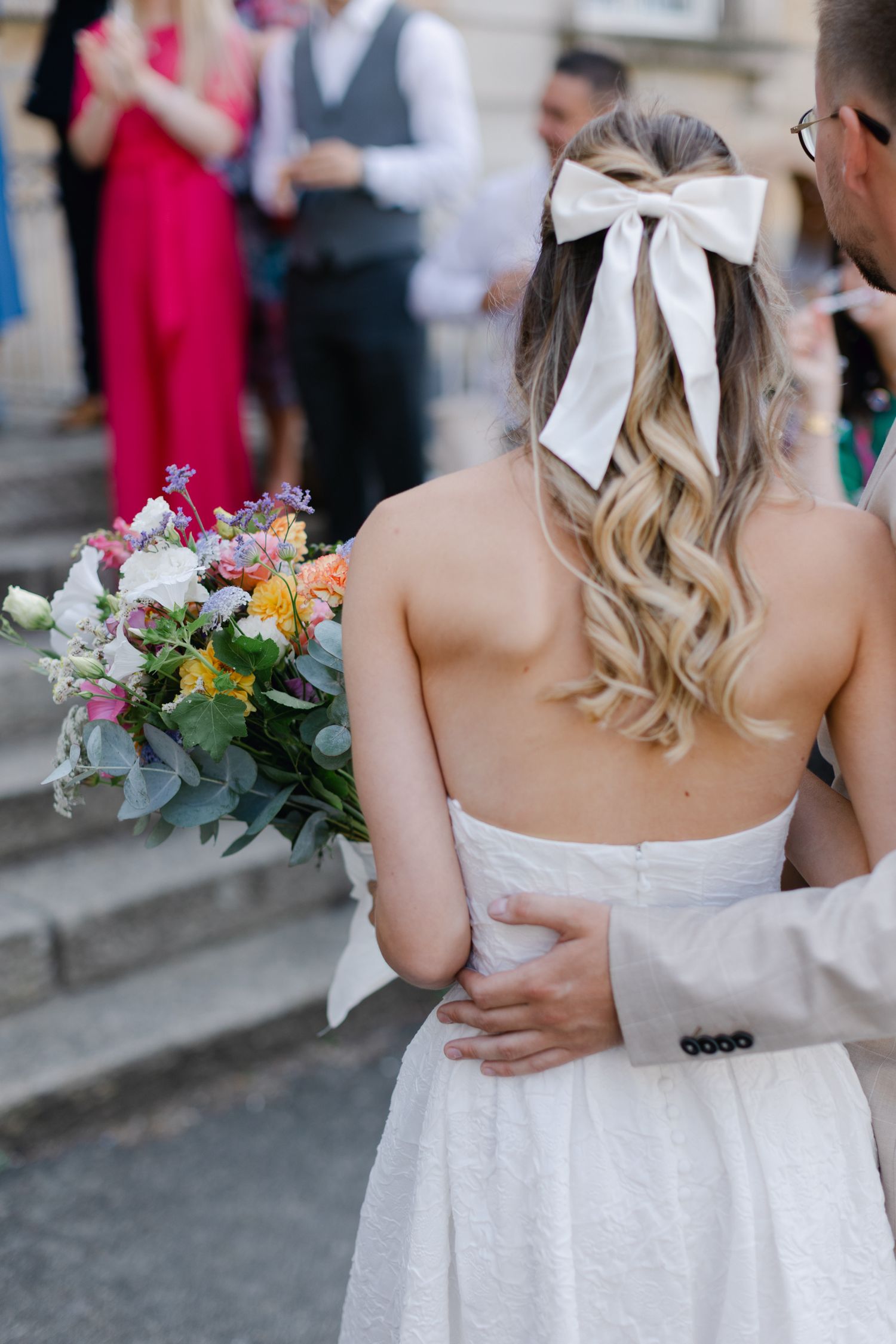 Vue arrière de la mariée avec un nœud blanc aux cheveux ondulés tenant un bouquet coloré, embrassée par le marié en costume beige.