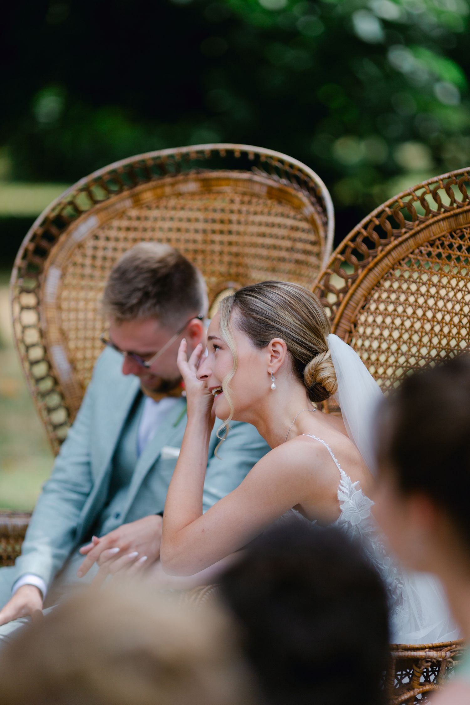 La mariée essuie ses larmes alors qu'elle est assise dans une chaise en osier à côté du marié lors de la cérémonie de mariage en plein air.