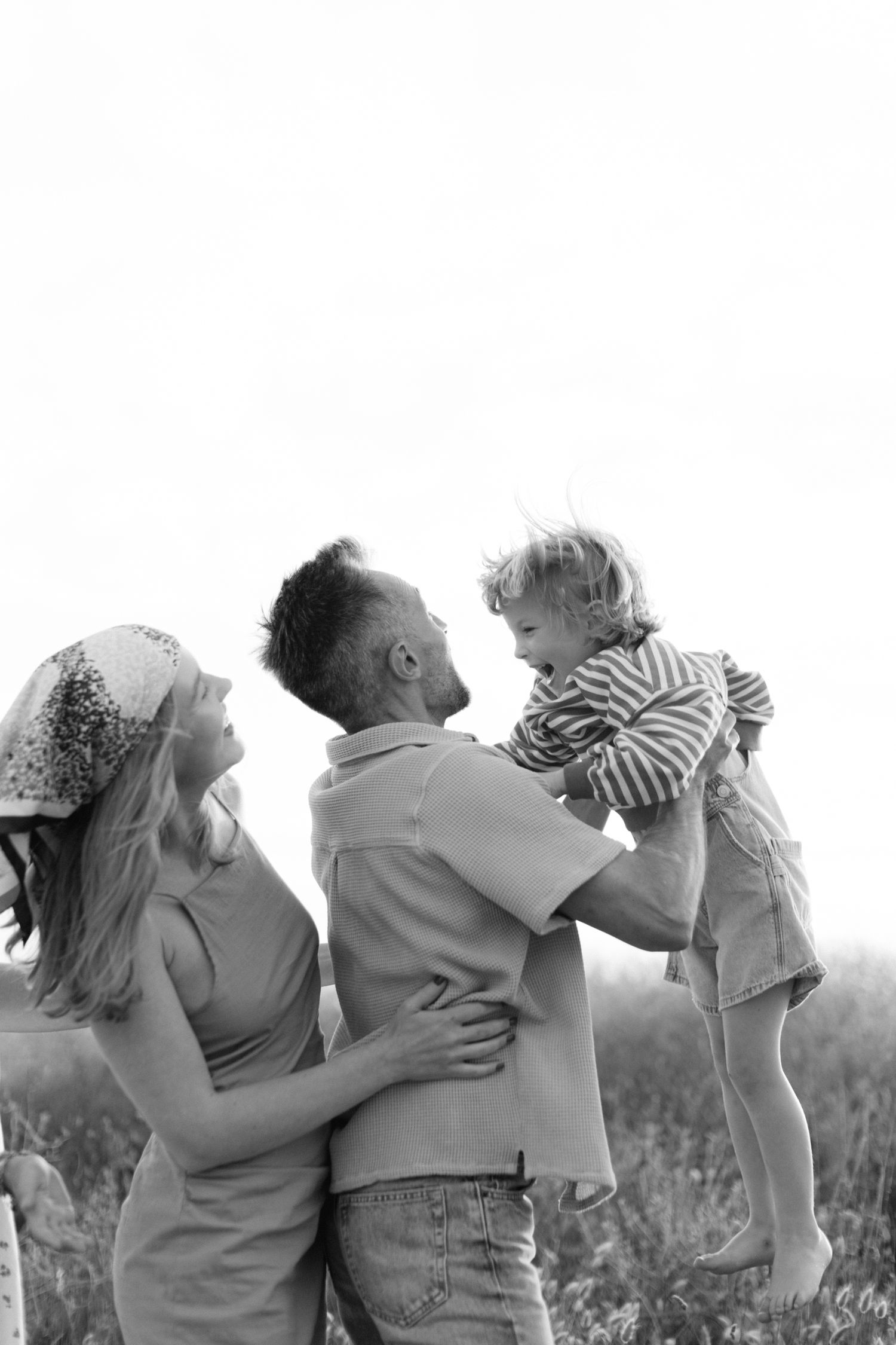 Photo en noir et blanc d'une famille à l'extérieur, père soulevant l'enfant tandis que la mère l'embrasse dans un champ.