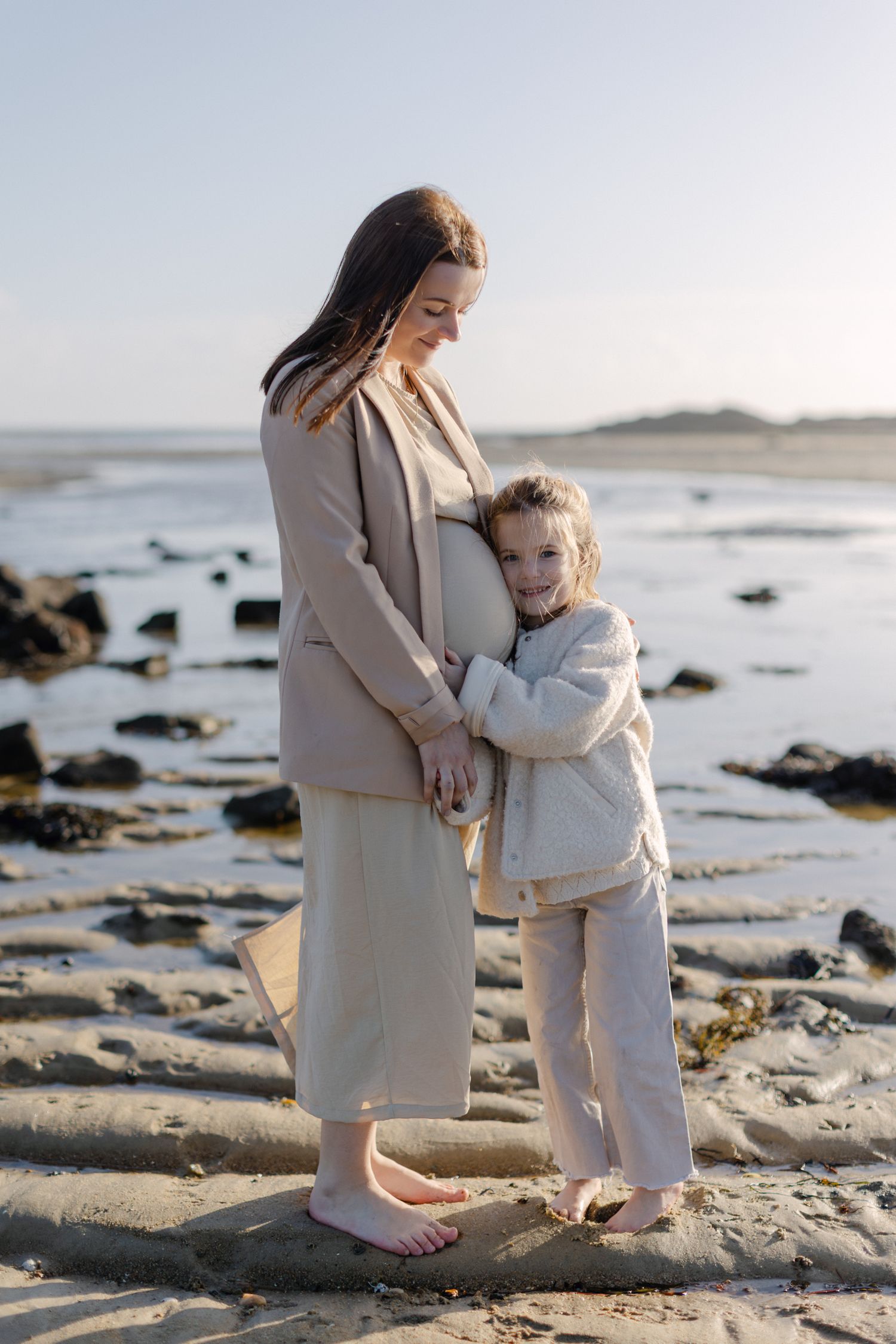 Une femme enceinte sourit alors que sa jeune fille embrasse sa bosse de bébé sur une plage de sable.