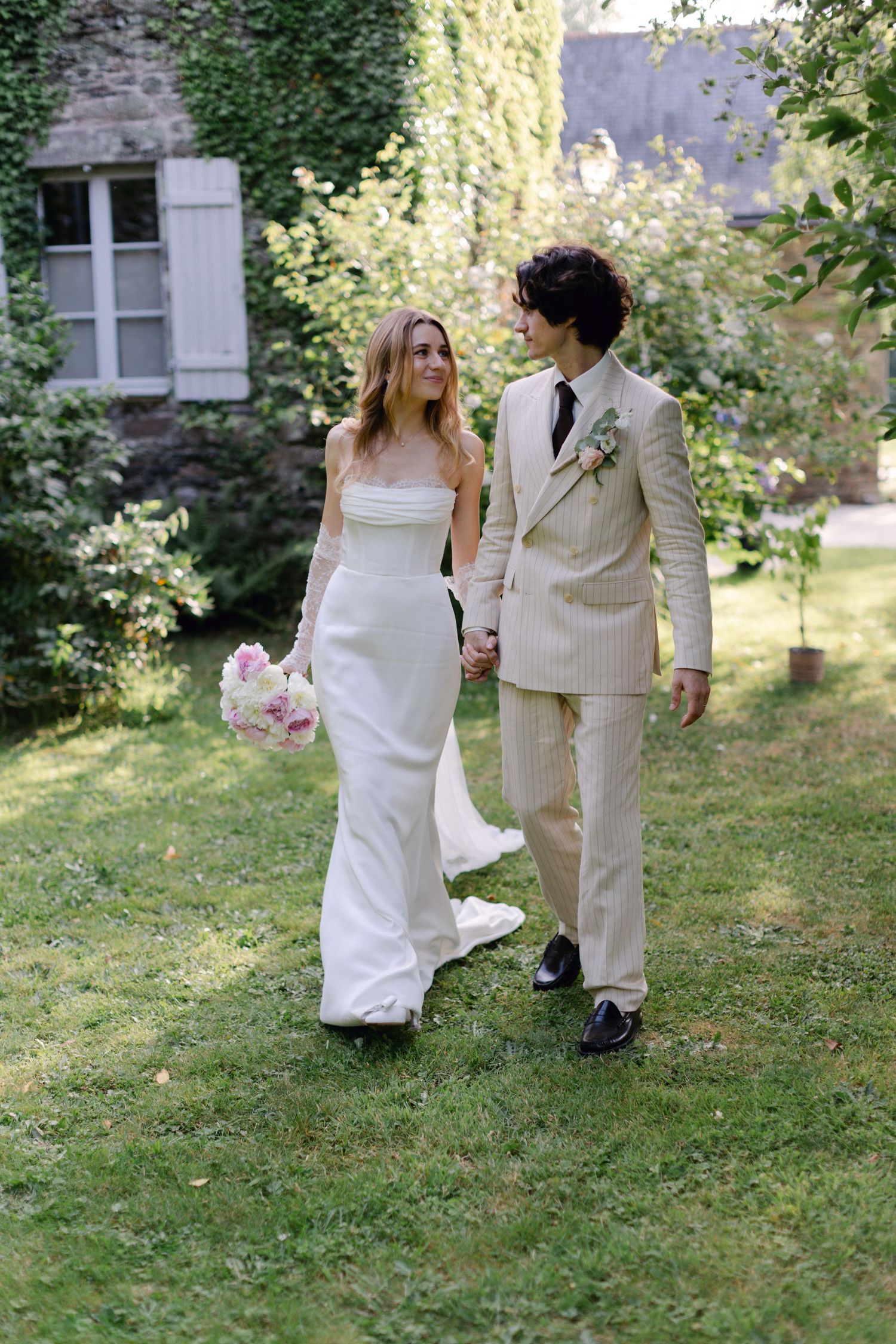 Les jeunes mariés marchent main dans la main à travers un jardin luxuriant, mariée en robe blanche, marié en costume à rayures beiges.