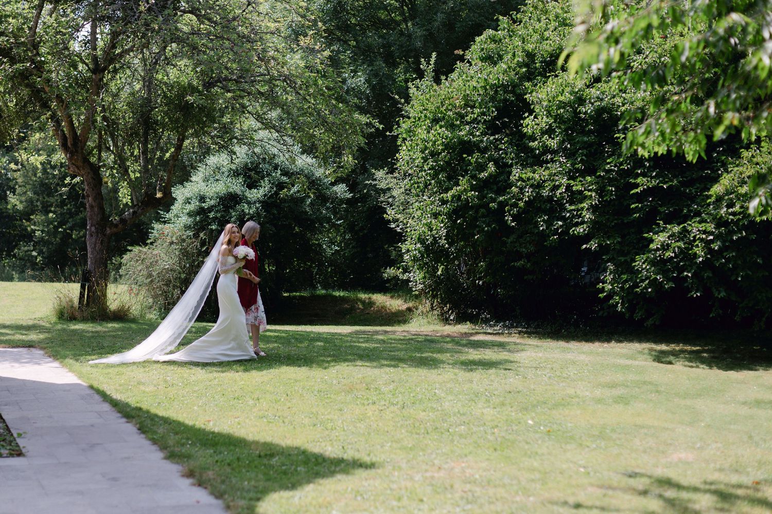 Mariée en robe de mariée blanche avec un long voile aux bras de sa mère dans un jardin vert luxuriant.