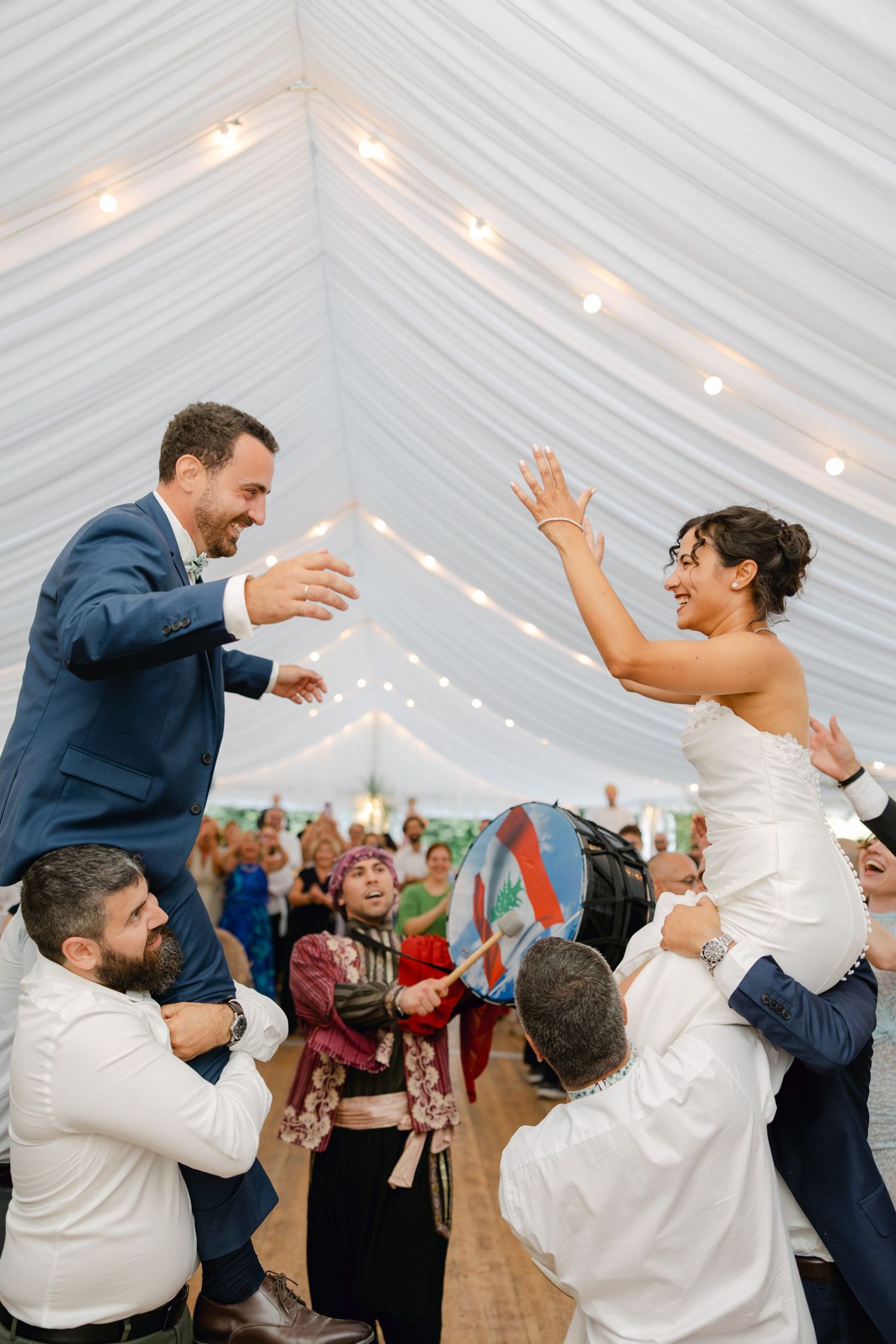 Bride and groom joyfully lifted by guests during lively wedding reception dance under a white tent with string lights.