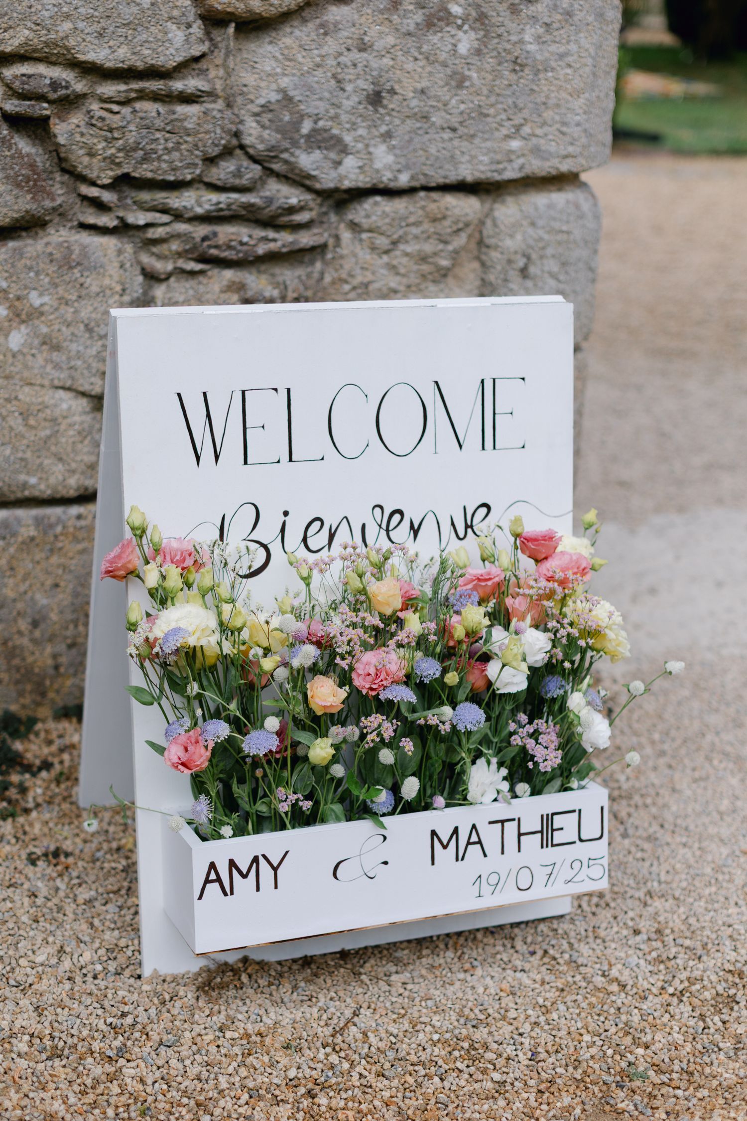 Panneau de mariage de bienvenue lisant Amy et Mathieu 19.07.25 décoré de fleurs fraîches colorées.