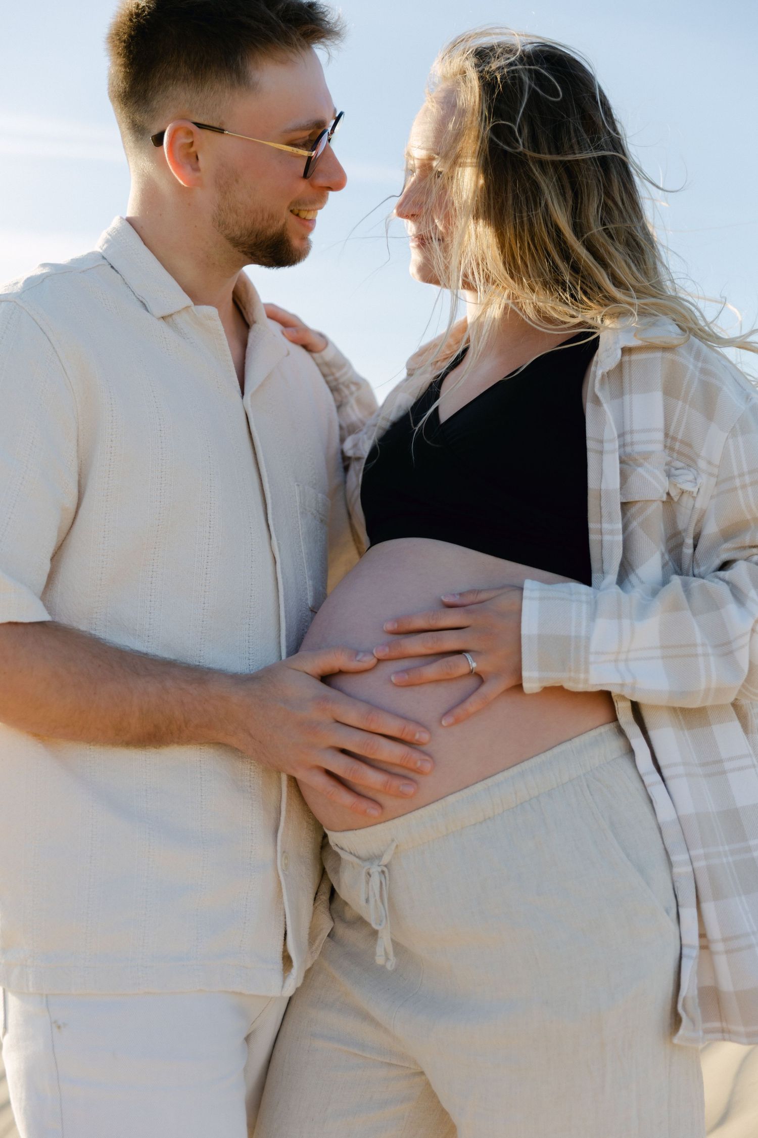 Couple souriant berçant le ventre de femme enceinte à l'extérieur, homme en chemise en lin, femme en haut à carreaux décontracté.