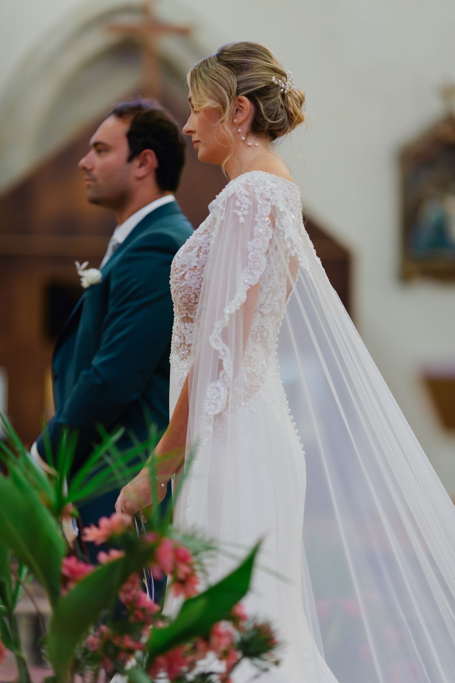 La mariée en robe de mariée cape en dentelle perlée se tient à côté du marié en costume sarcelle lors de la cérémonie à l'église.