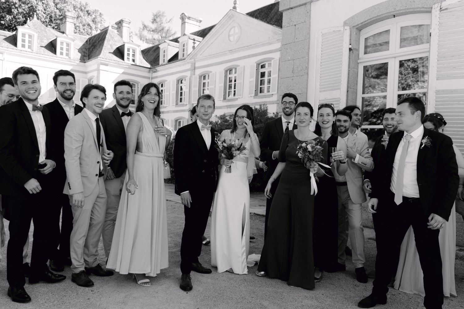 Photo de mariage en noir et blanc de la mariée, du marié et des invités posant à l'extérieur d'un élégant domaine.