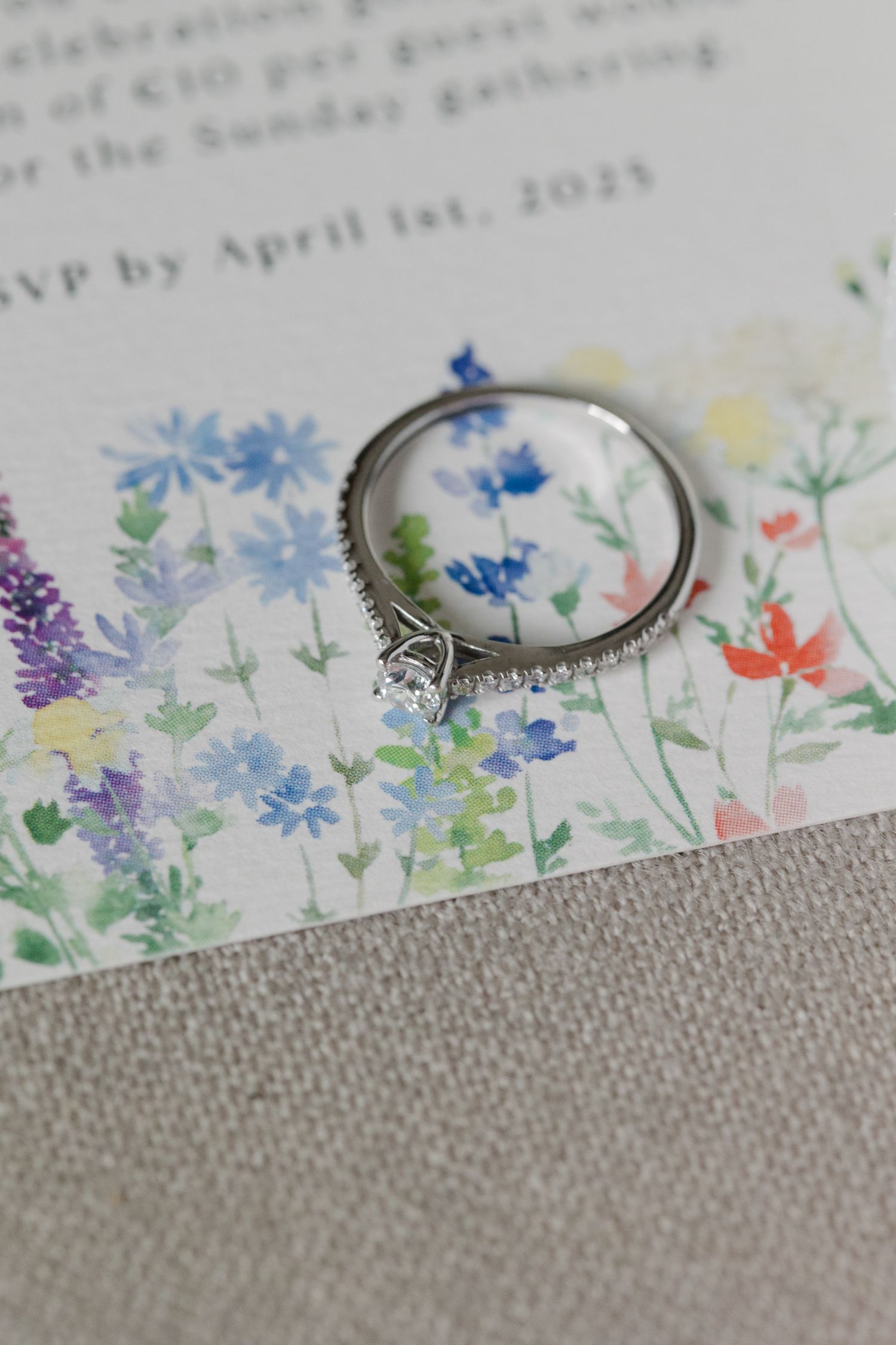 Bague de fiançailles solitaire en diamant avec bande pavé reposant sur une invitation de mariage à l'aquarelle florale.