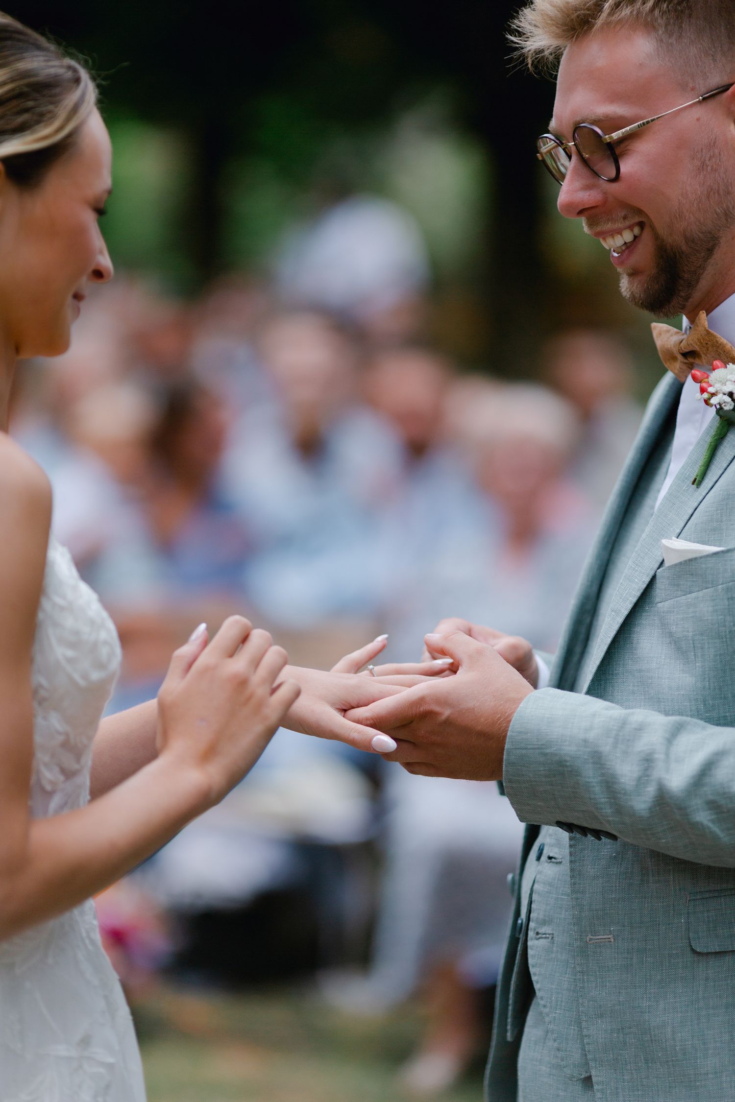Marié souriant en costume vert sauge recevant une bague de mariage de la mariée lors d'une cérémonie en plein air.