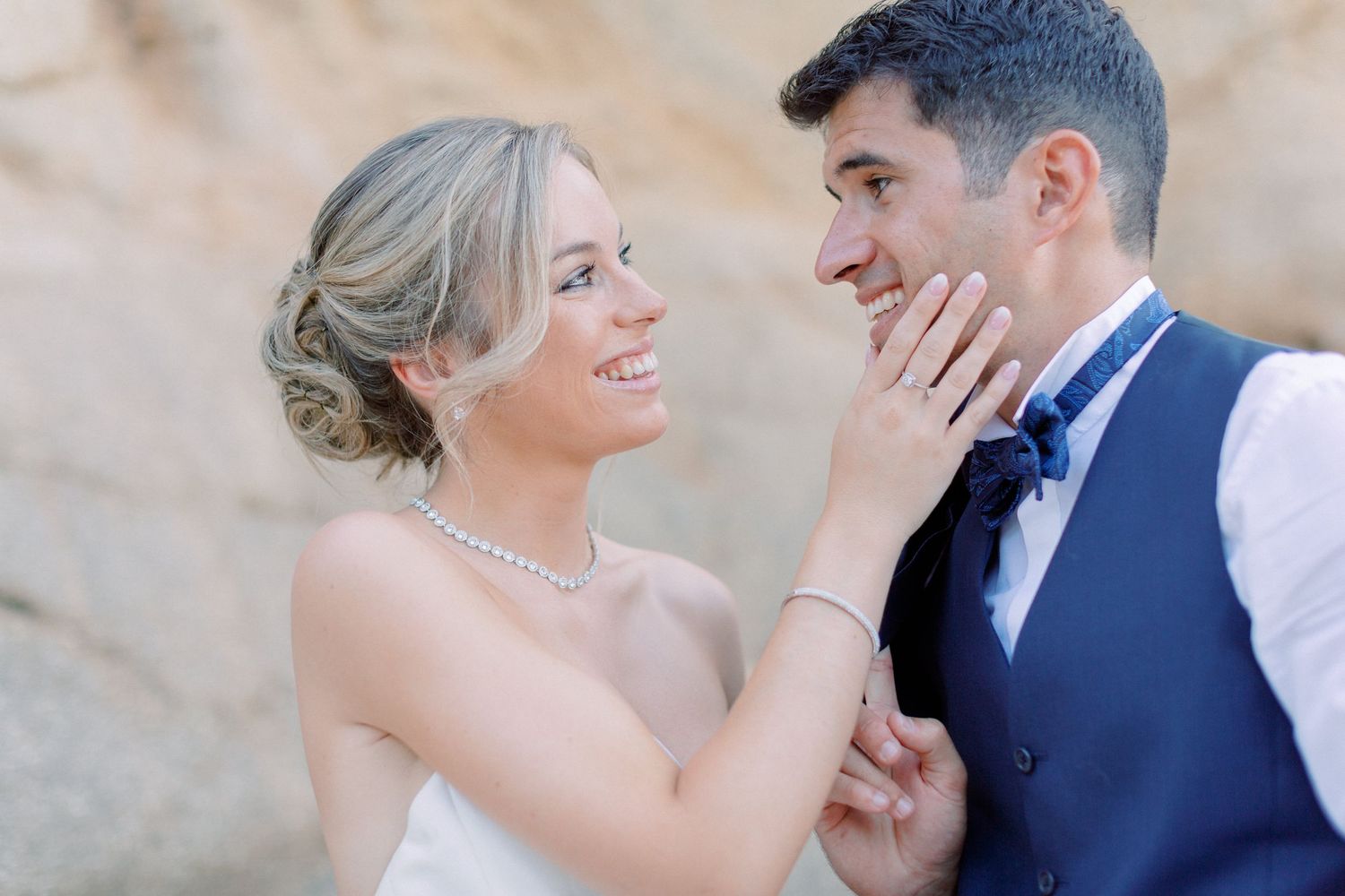 Une mariée souriante vêtue d'une robe blanche et d'un collier de perles touche tendrement la joue de son marié dans un gilet bleu marine.
