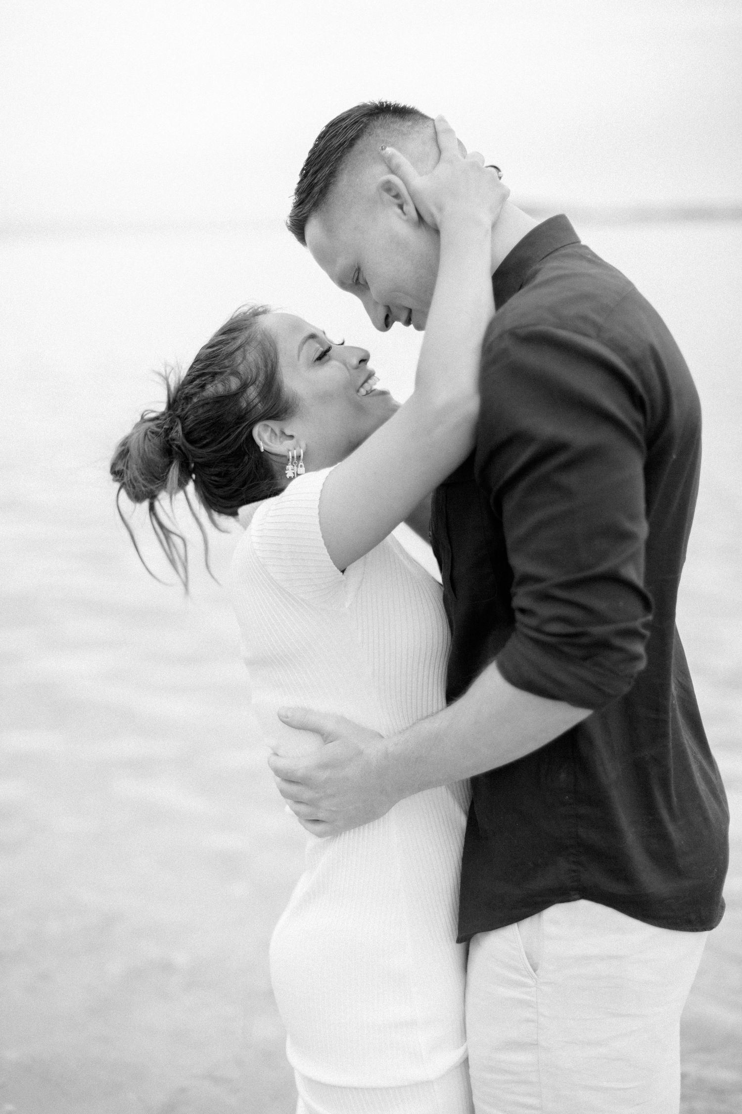 Photo en noir et blanc d'un couple s'embrassant et s'embrassant presque sur une plage, femme en robe blanche, homme en chemise sombre.
