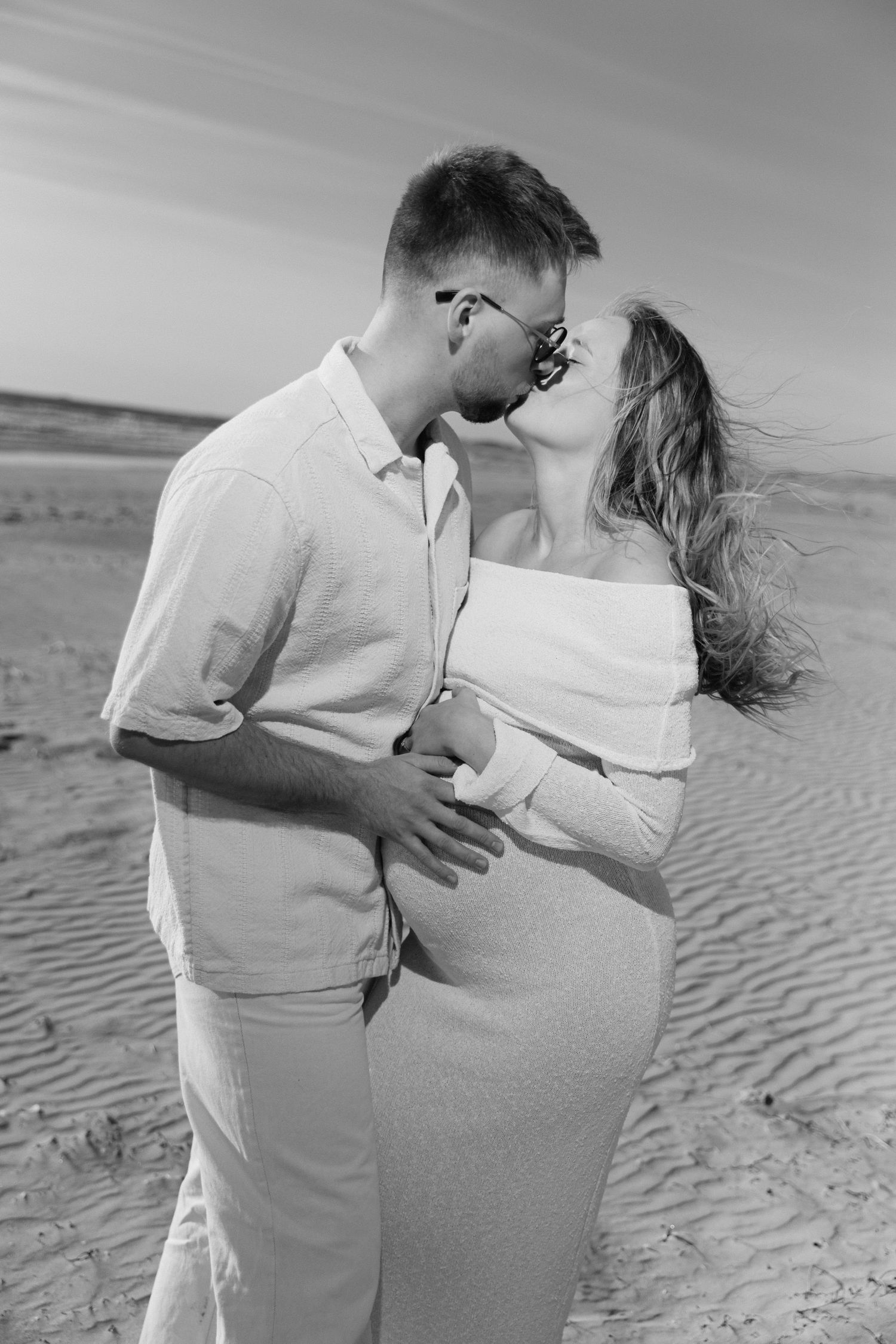 Photo en noir et blanc d'un couple enceinte s'embrassant sur une plage de sable, femme en robe blanche.
