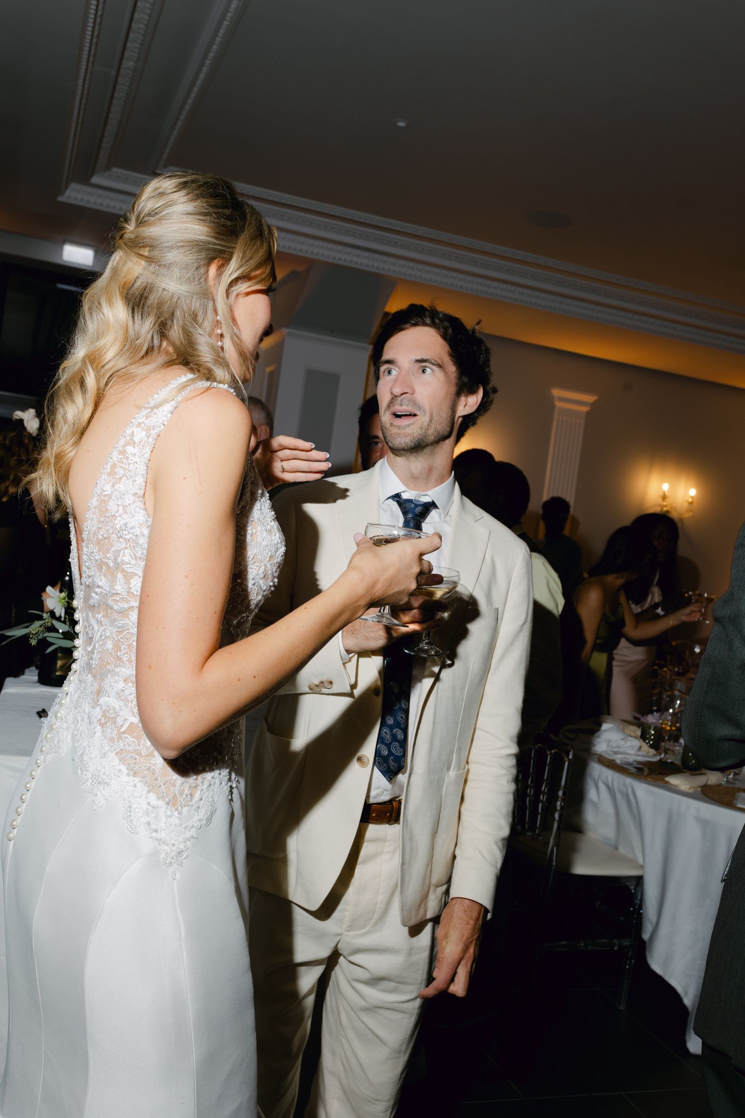 Mariée en robe dos nu en dentelle discutant avec un invité masculin en costume crème lors d'une élégante réception de mariage.