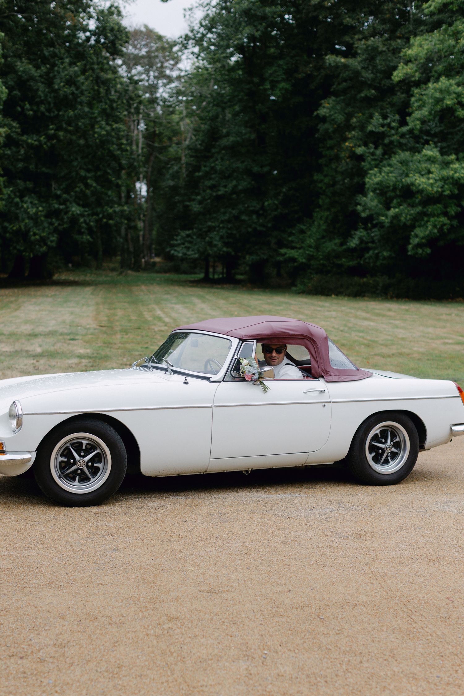 Voiture MG convertible vintage avec toit bordeaux garé sur une allée de gravier entourée d'arbres.