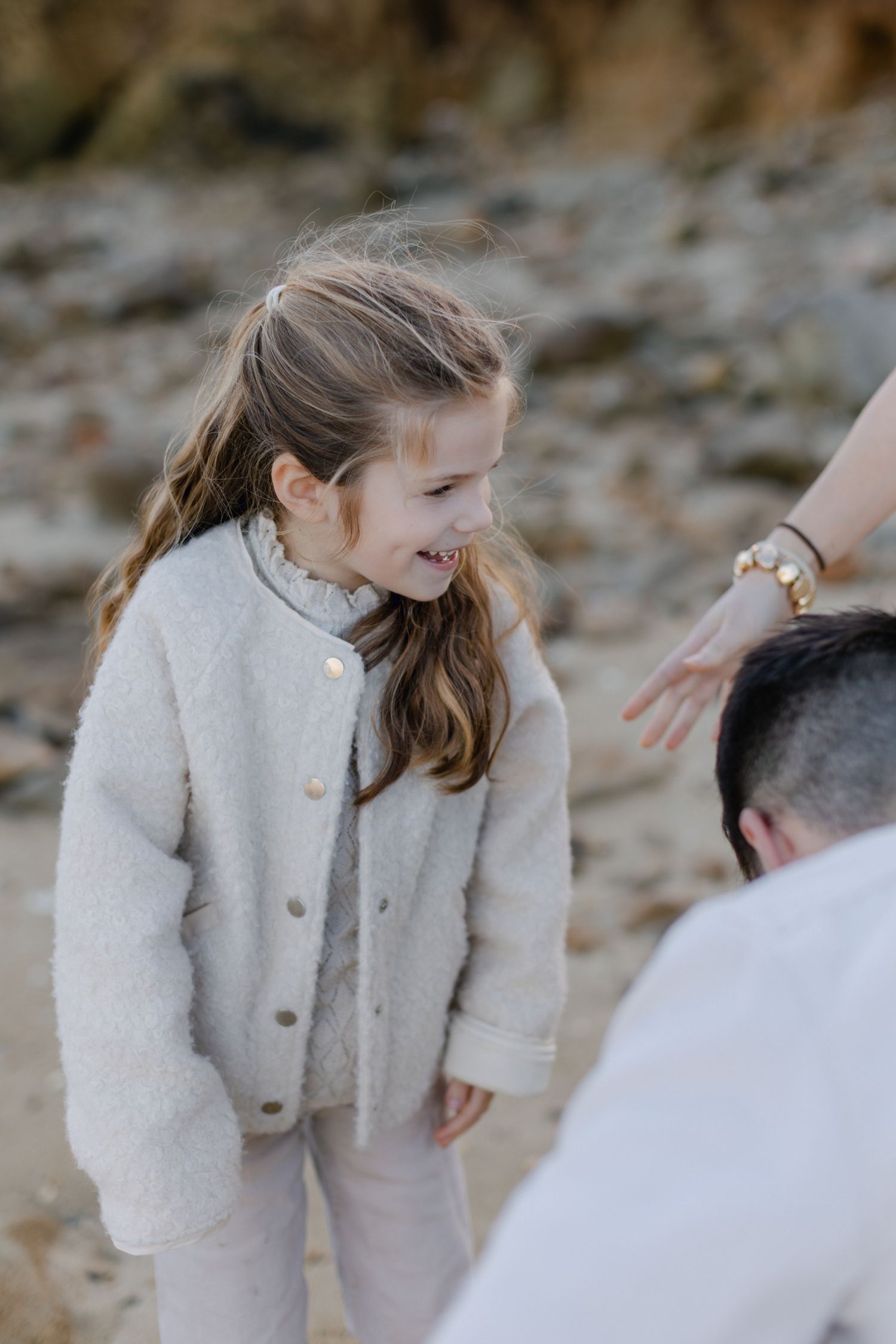 Une jeune fille heureuse en veste crème sourit et tend la main sur une plage rocheuse.