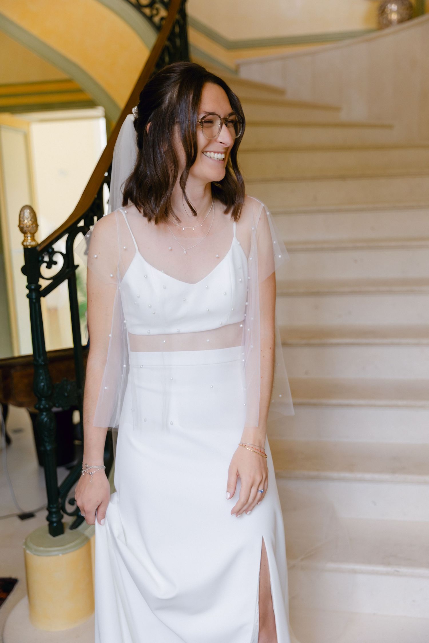 Mariée souriante en robe blanche à bretelles spaghetti avec superposition transparente et voile, debout sur un élégant escalier.