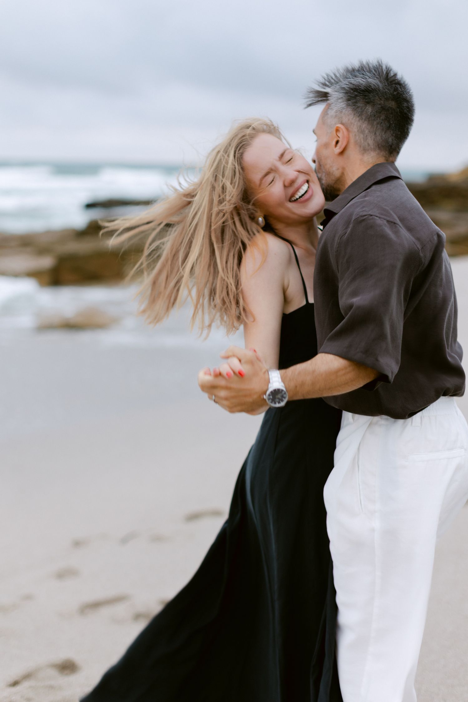 Couple riant et dansant ensemble sur une plage de sable, femme en robe noire avec les cheveux qui soufflent dans le vent.