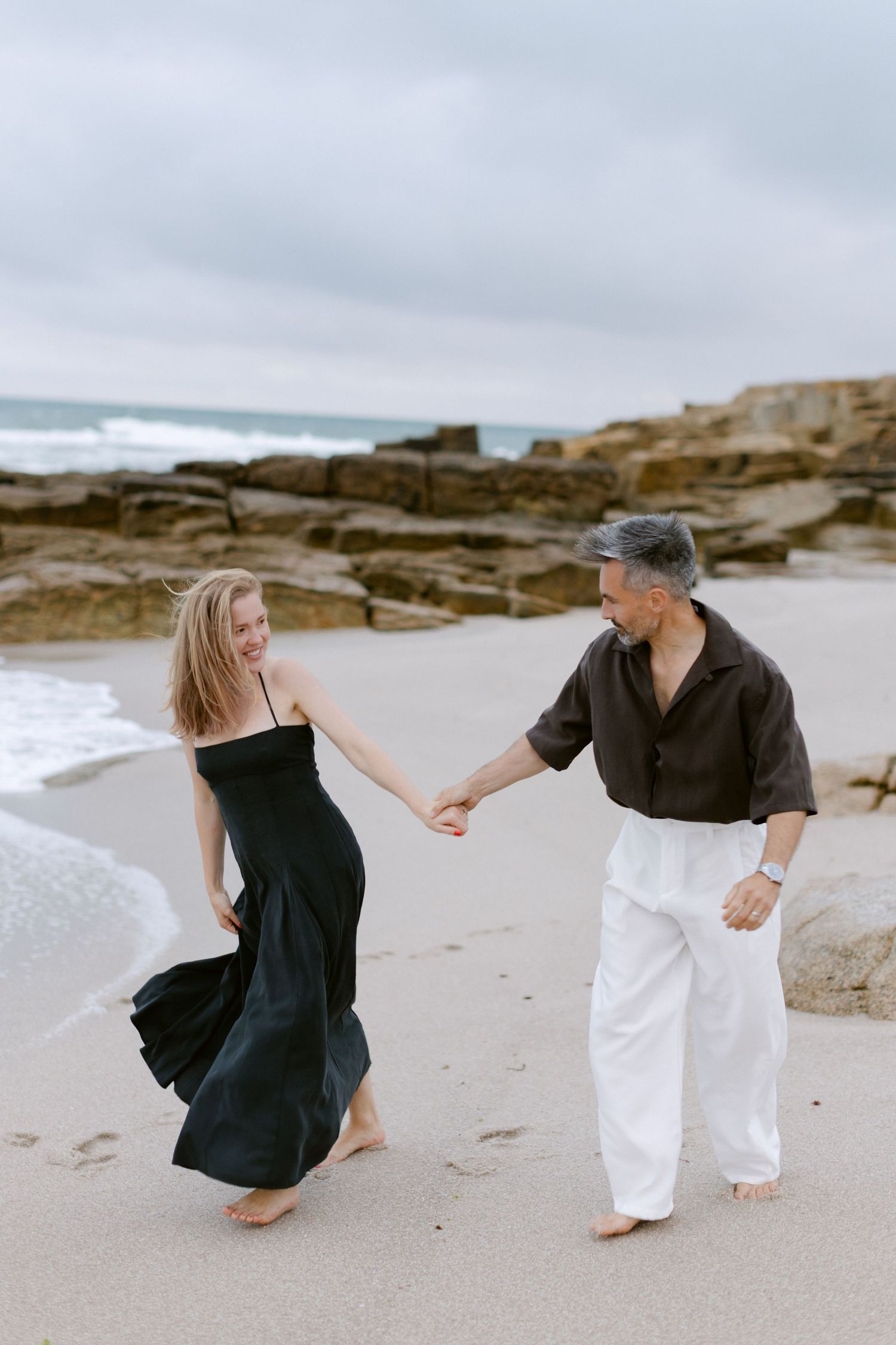A couple holds hands and dances barefoot on a sandy beach, woman in black dress, man in dark shirt and white pants.
