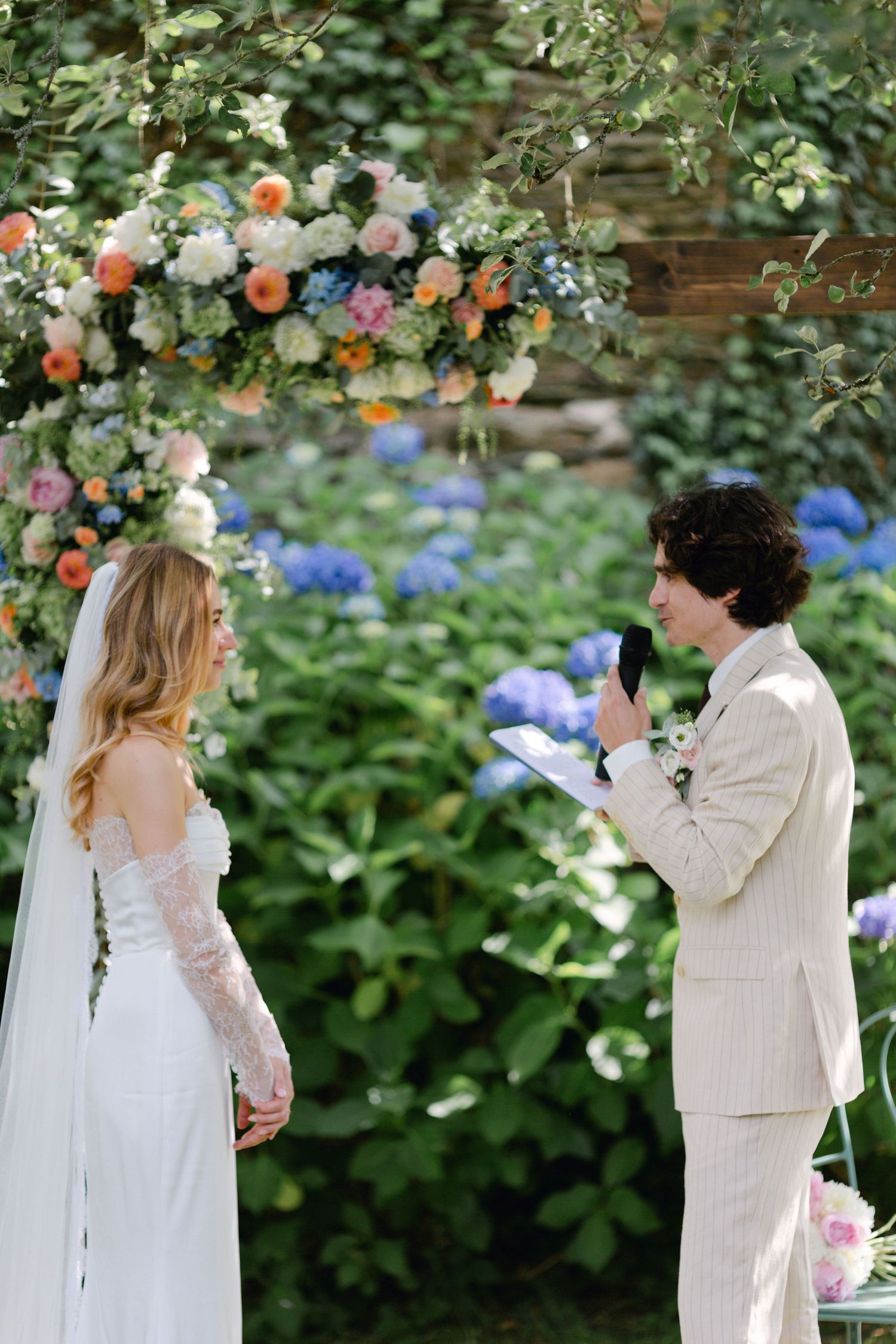 Le marié en costume beige lit les vœux de la mariée lors d'un mariage dans le jardin en plein air sous une arche florale colorée.
