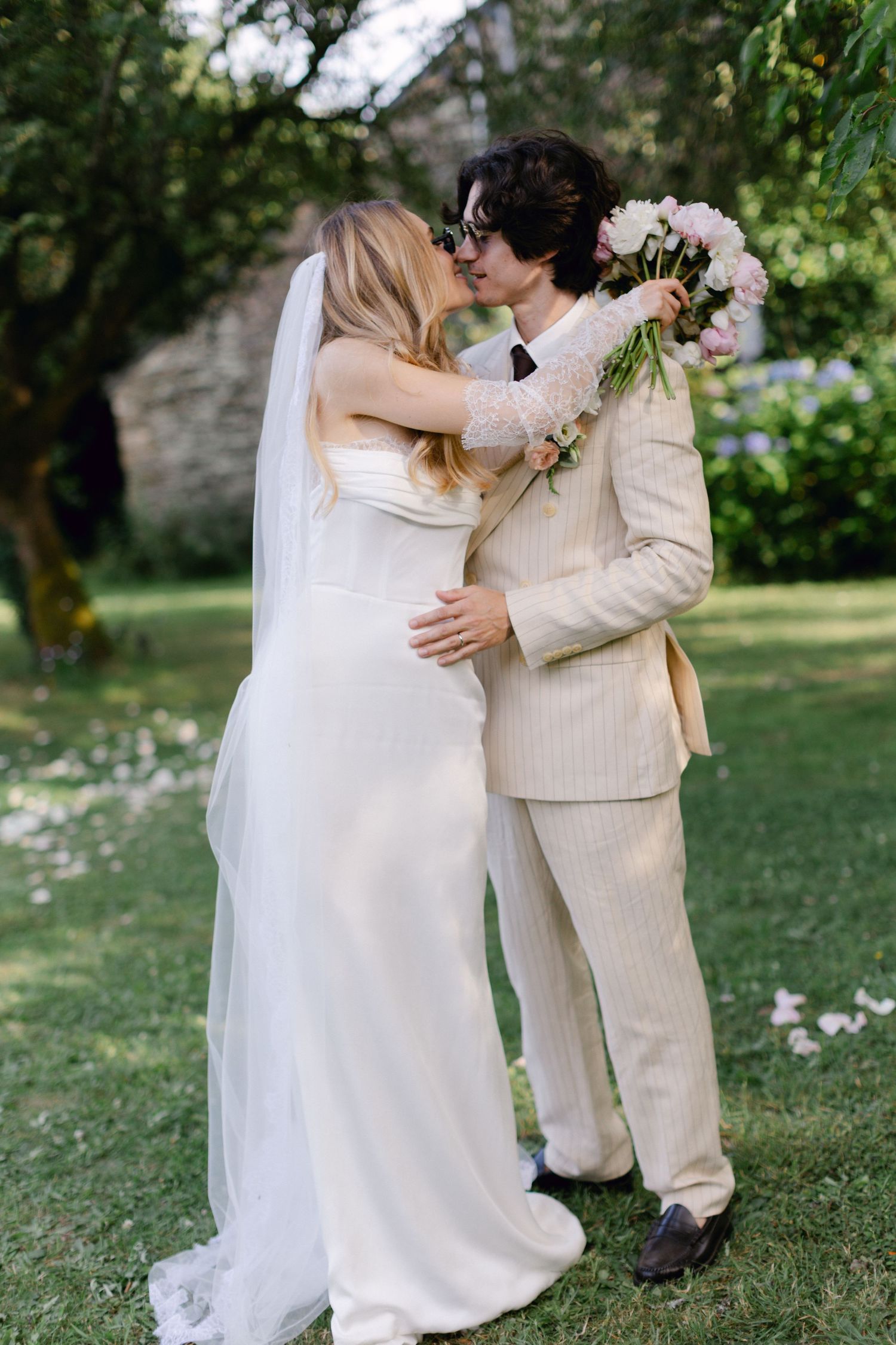 La mariée et le marié partagent un baiser romantique sur la pelouse du jardin, elle en robe blanche avec voile, lui en costume à rayures fin